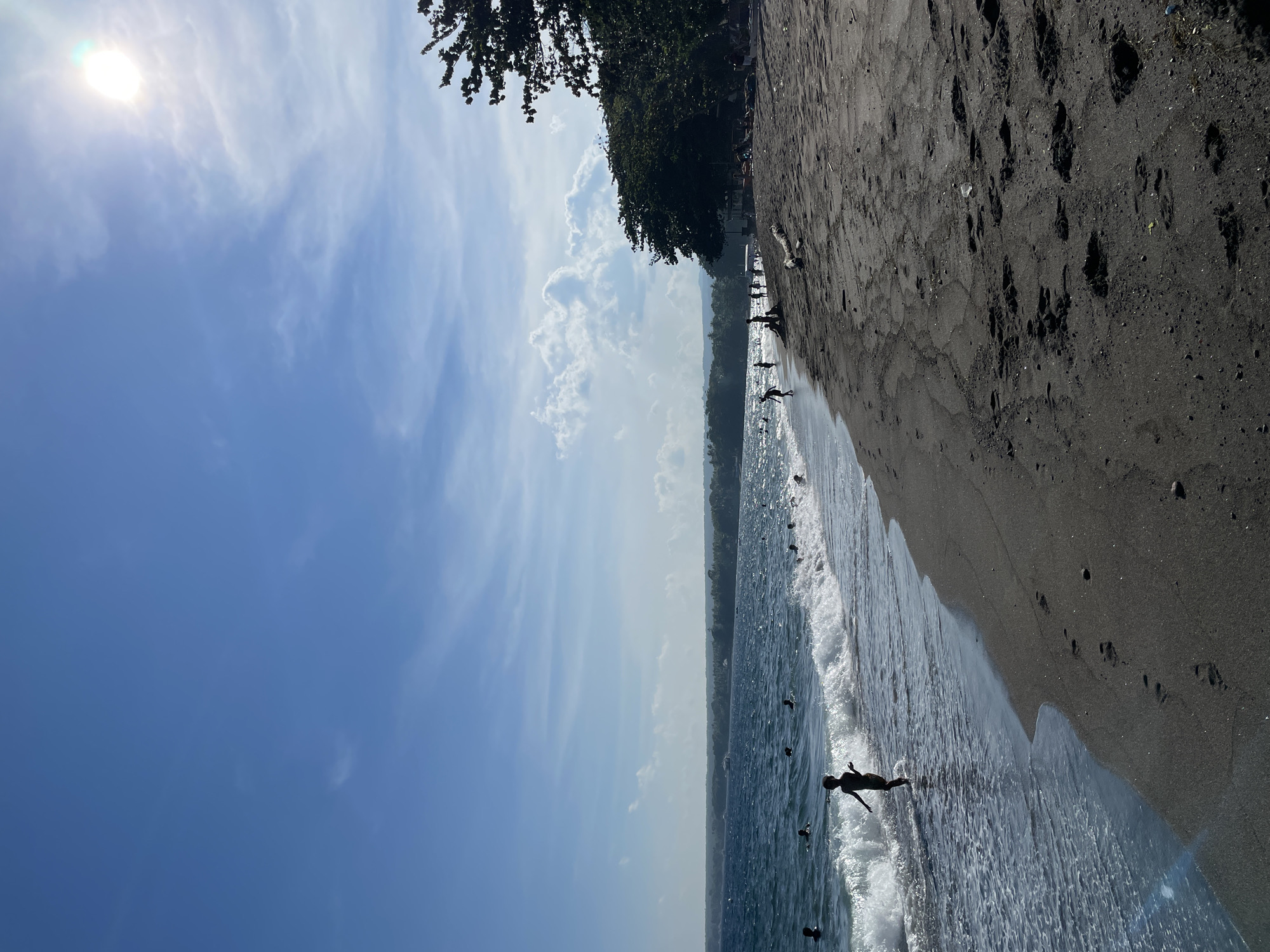 This image depicts a sunny day at a beach in Dauin, Philippines. The sea is a deep blue with moderate waves, and several people are visible swimming or wading in the water. The shoreline is made of dark sand, with footprints clearly visible. To the right, lush green trees line the edge of the beach, and in the distance, a faint outline of land can be seen across the water. The sky is a vibrant blue with scattered white clouds and the sun is bright, casting a glare in the upper right portion of the image.