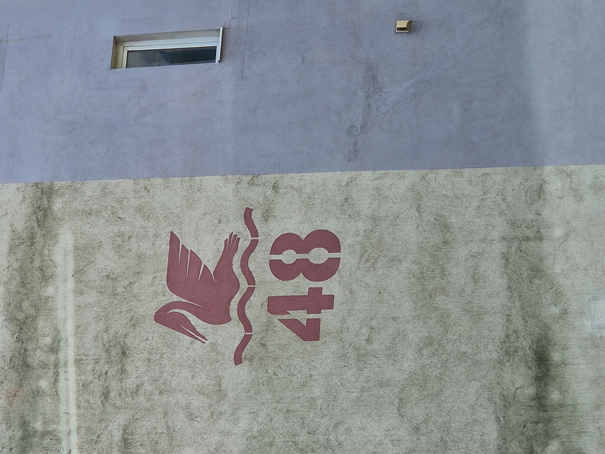 The image captures a section of a building's exterior wall, divided horizontally into two distinct painted areas. The upper half of the wall is a solid, light purplish-gray color with a slightly mottled texture. Towards the top left of this section, a small, rectangular window with a white frame is visible, set into the wall. Further to the right, near the top edge, a small, square, gold-colored or brass-colored fixture protrudes from the wall.
The lower half of the wall features a textured, light beige or off-white surface, which appears somewhat aged or weathered. Dominating this lower section is a large, stylized graphic rendered in a muted reddish-pink or maroon color. On the left, a bird, possibly a pelican or a similar water bird, is depicted in profile, facing left, with a wavy line beneath its body, suggesting water. To the right of the bird, the numbers "48" are stenciled vertically in the same reddish-pink color. The numbers are stylized with segmented gaps, characteristic of stenciling. The overall impression is a decorative element on a building facade.
There are no people or specific activities depicted. The lighting suggests it is daytime, but no strong shadows indicate a precise time. The setting is an urban or residential building exterior.
