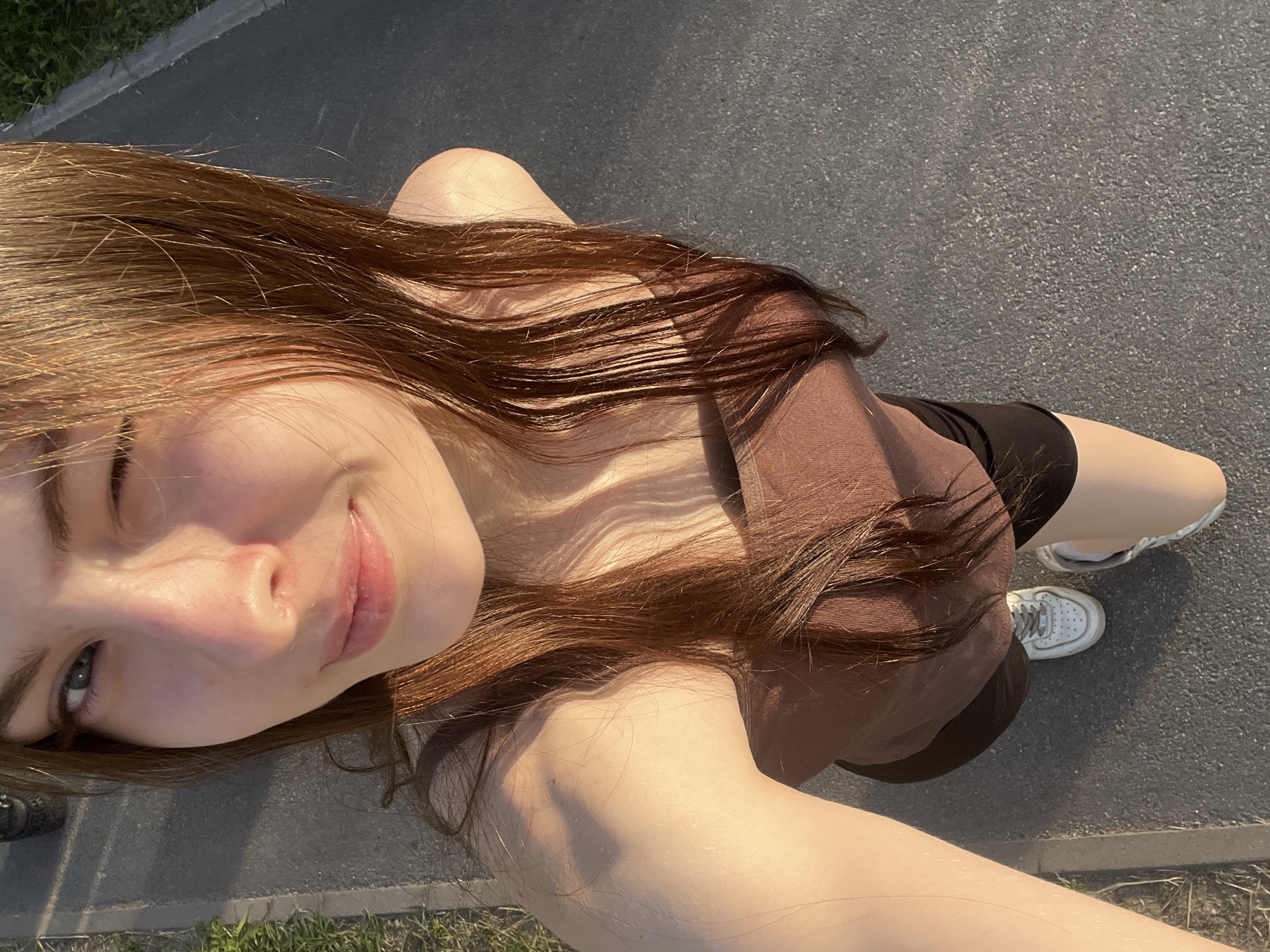 A young female is captured in a high-angle selfie photograph, standing outdoors on an asphalt paved path in Saint Petersburg, Russia. She has long, straight brown hair, illuminated by golden hour sunlight, falling over her shoulders and chest. She is winking with her right eye closed and her left eye open, displaying a subtle smile. Her skin appears fair with a warm, reddish tint on her nose and cheeks, likely from the direct sunlight. She is wearing a brown strapless tube top and black shorts. White athletic shoes are visible on her feet. To the left of the paved path, a narrow strip of green grass and dirt is present. The sun casts distinct, elongated shadows to the right and behind the subject, indicating a low sun angle.