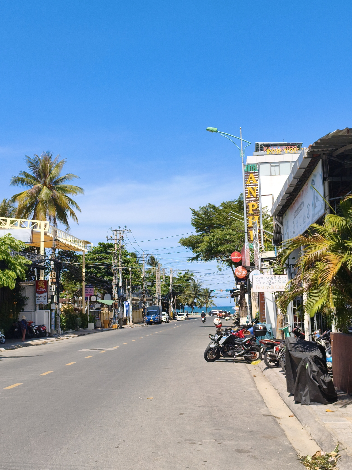 The image captures a street scene in Nha Trang, Vietnam, on a clear, sunny day. The street runs lengthwise, paved and marked with yellow dashed lines, with a sidewalk on the right side. Several parked motorcycles are visible on the sidewalk, some covered with black tarps. A blue delivery van is driving down the street, and other vehicles are present in the distance. Buildings line the street, featuring signage for "HOTEL AN PHI" and "KOKORO Bistro & Cafe." Palm trees and other foliage are interspersed with the buildings, and power lines stretch across the scene. The sky is a vibrant blue, with a hint of the ocean visible in the background at the end of the street.