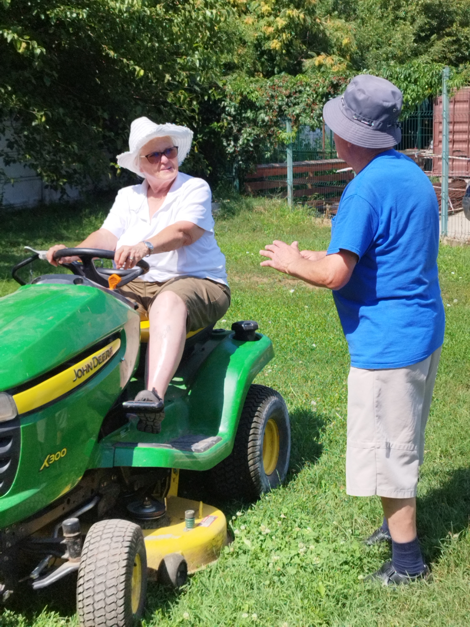 An outdoor, full-shot, eye-level photograph captures a bright, sunny day in Bucharest, Romania. In the foreground, a woman is seated on a green John Deere lawn tractor, wearing a white sun hat and glasses, with her hands on the steering wheel. She is dressed in a white polo shirt and khaki shorts. To the right of the tractor, a man stands facing the woman, wearing a blue t-shirt and light-colored shorts, with his hands raised in a gesture of conversation. He is wearing a grey bucket hat. The background consists of a lush green lawn, trees, and a fenced area with structures, suggesting a residential or park-like setting. The overall scene depicts a casual interaction between two individuals on a warm day.