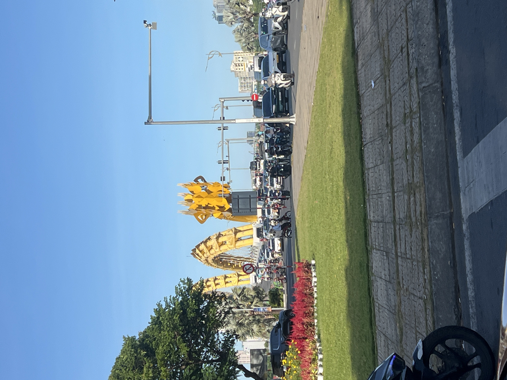 The image captures a view of the Dragon Bridge in Da Nang, Vietnam, from street level. The iconic golden dragon structure is prominently featured in the background, spanning across a busy intersection. A mix of cars and motorbikes are navigating the road, indicating moderate traffic. In the foreground, a well-maintained green lawn is bordered by a vibrant display of red flowers in white planters. The sky is clear and blue, suggesting a sunny day. The environment appears urban, with buildings and palm trees visible in the distance. The overall scene depicts a typical day on a major road in Da Nang, highlighting its famous landmark.