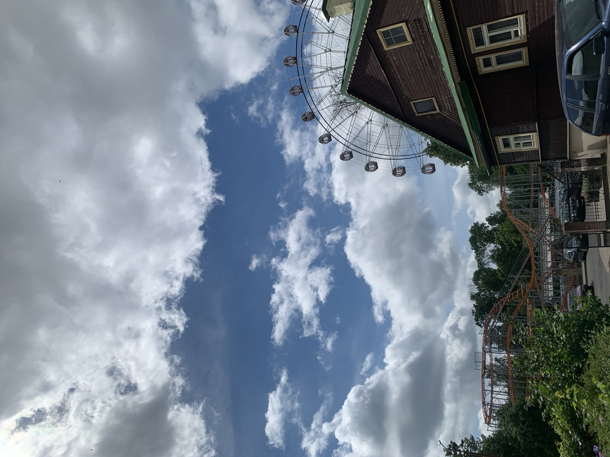 This portrait-oriented image captures an upward view of an amusement park in Hrodna, Belarus, under a partly cloudy sky during the daytime. The frame is largely filled with a mix of bright blue sky and white and grey cumulus clouds, with small dark specks, possibly birds, visible against the clouds in the upper left.
On the right side, a large, light-colored Ferris wheel with multiple cabins extends from the top to near the bottom of the frame. Adjacent to it, a two-story building with a dark brown wooden facade and a green roof trim is visible, featuring several windows with light-colored frames. A dark blue car is partially parked near this building on the far right.
In the lower right, an orange roller coaster track with grey supports is partially visible, weaving through dense green trees and bushes that also border the bottom of the image. No specific activity or visible text is present in this static scene of the park's infrastructure.