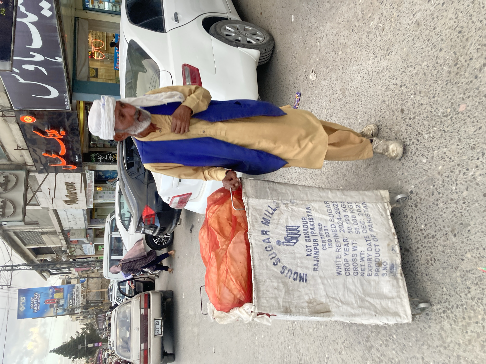 An outdoor scene in Quetta City Tehsil, Pakistan, depicts a man pulling a cart down a street. The man is dressed in traditional attire consisting of a beige tunic and pants, a white turban, and a blue scarf draped over his shoulders. He is seen holding onto a metal handle attached to a white sack. The sack is labeled "INDUS SUGAR MILLS" and contains details such as "KOT BAHADUR RAJANPUR (PAKISTAN)", "WHITE REFINED SUGAR", "CROP YEAR: 2024-2025", "GROSS WT: 50.105 KGS", "NET WT: 50.000 KGS", "EXPIRY DATE DEC: 2027", and "PRODUCT OF PAKISTAN". Inside the sack is an orange mesh material, likely containing goods. The man is walking on a concrete road alongside parked cars, including a white car with its rear end visible and a beige car. In the background, there are shops with signs written in Urdu and a large advertisement banner for "SMART HEATING" on a street with heavy traffic. Overhead power lines and poles contribute to the urban environment.