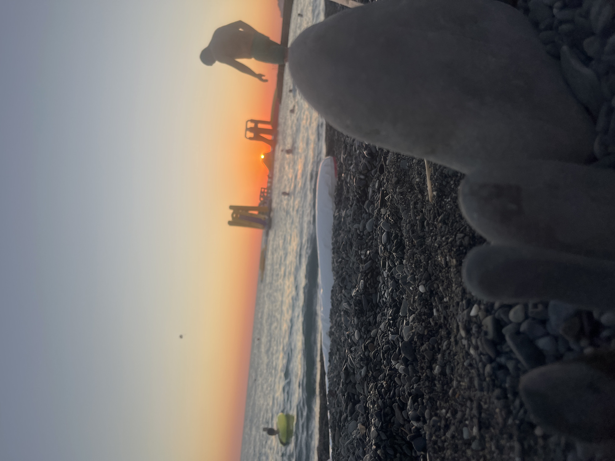 The image depicts a sunset scene on a pebbled beach in Sochi, Russia. In the foreground, out-of-focus toes and rocks are visible. The middle ground shows the sea with waves, people swimming and on inflatable rings, and water slides. A silhouette of a person is on the right side of the frame. The background features a vibrant sunset with the sun low on the horizon, casting a warm glow over the water and sky.
