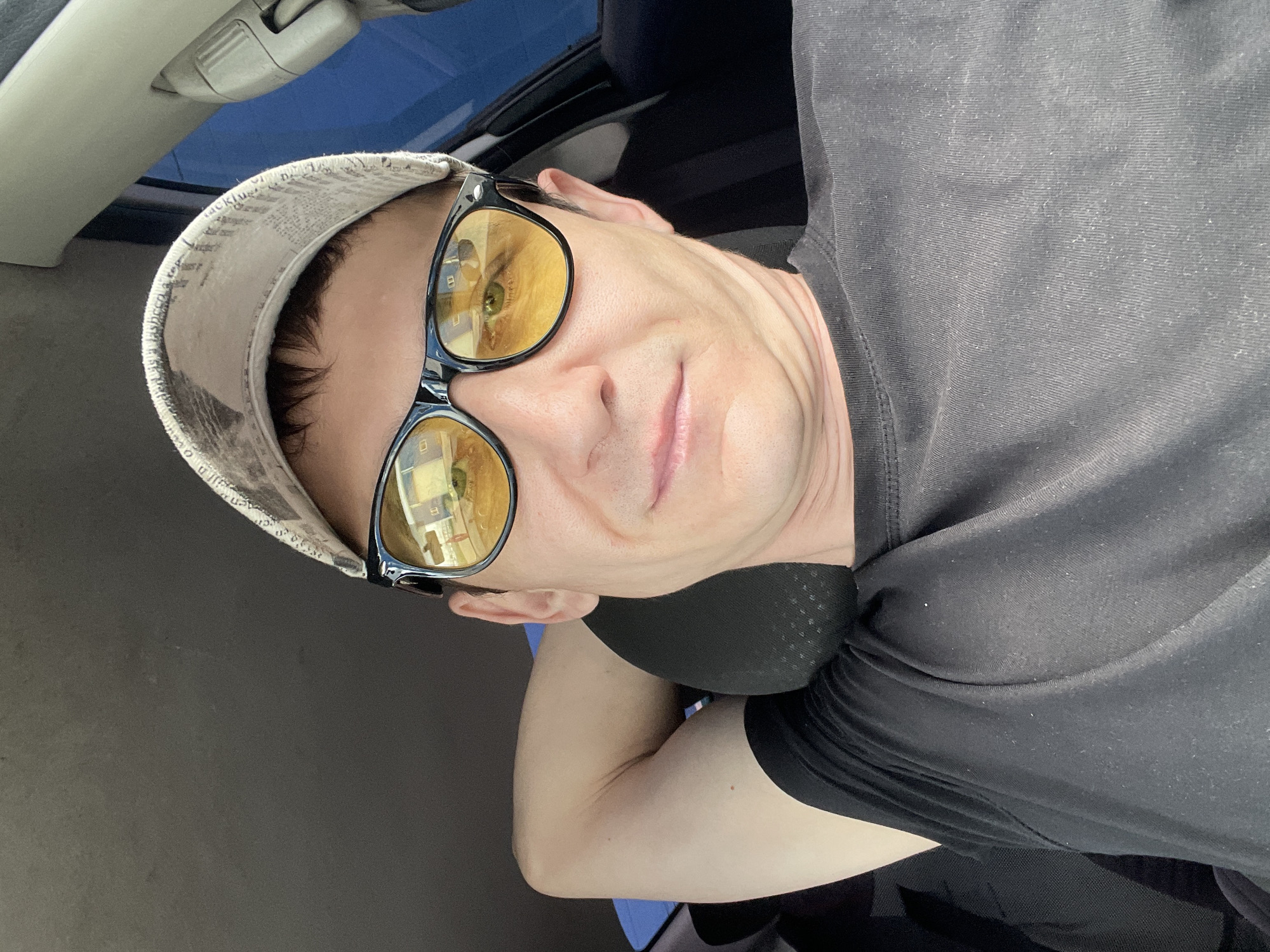 A man wearing sunglasses and a cap is sitting in the driver's seat of a car. He has his arm behind his head and appears to be looking at the camera. The reflection in his sunglasses shows a building and a street, suggesting he is in a city environment. The interior of the car is visible, including the headrest and part of the dashboard. The scene is likely taking place during the daytime, as indicated by the lighting.