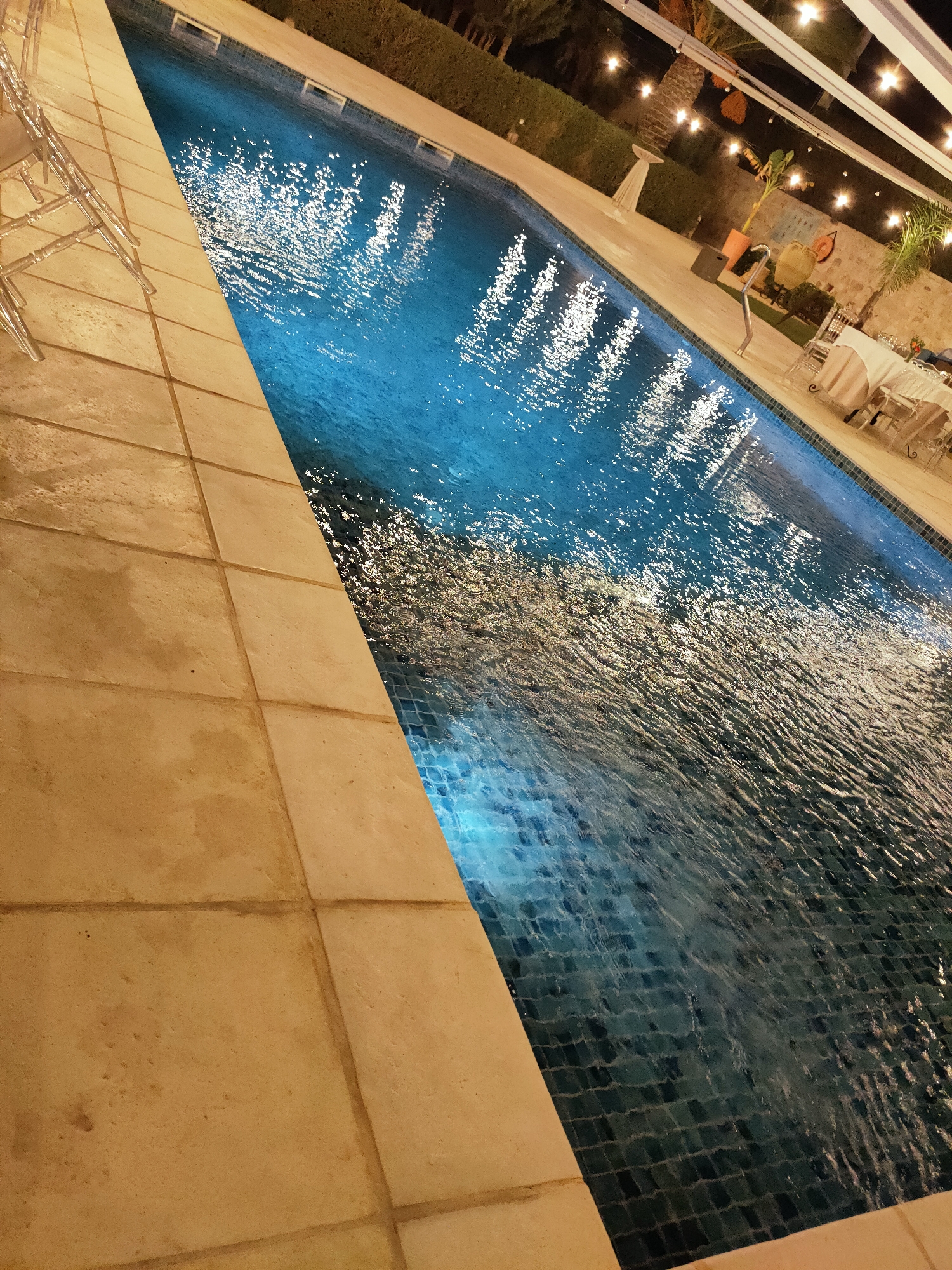 An outdoor swimming pool is illuminated at night, reflecting lights from above and surrounding string lights. The pool water is a deep blue, with visible ripples and light reflections on its surface. The pool is bordered by tan-colored stone tiles, which are wet in places. To the left of the pool, several clear plastic chairs are visible. In the background, there is a low hedge, a palm tree, and a stone wall. A white tent-like structure with string lights hangs overhead. A table covered with a white tablecloth and chairs is set up near the pool in the background, suggesting a dining or event setting. The scene appears to be a resort or a private residence in Cebalat, Tunisia.