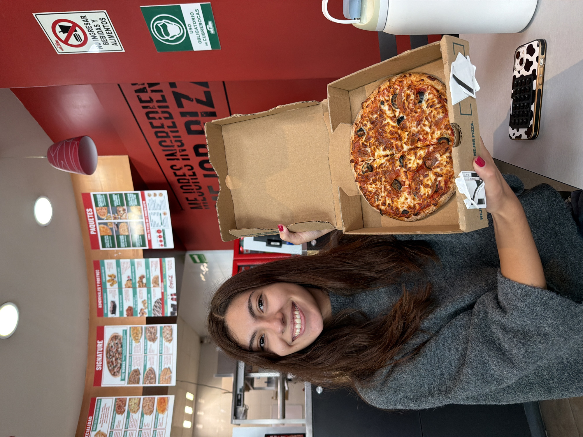 A young adult female, appearing to be in her 20s with long dark brown hair and brown eyes, smiles broadly at the camera while holding an open small-sized pizza box containing a pepperoni and black olive pizza. She wears a grey, textured, long-sleeved sweater. Her left hand, with red painted fingernails, grips the side of the cardboard pizza box. The pizza inside is cut into six slices. On the light grey tabletop to her right, a white and light blue water bottle stands next to a smartphone in a cow-print case, placed face down.
The scene is an indoor restaurant, identified as a Papa John's in San Bernardino Tlaxcalancingo, Mexico, based on branding elements. A prominent red wall in the background displays large black lettering that reads "MEJORES INGREDIENTES MEJOR PIZZA" (Better Ingredients. Better Pizza.). Below this, to the right, are two official-looking signs: a white sign with a red circular 'no' symbol indicating "NO INGRESAR BEBIDAS Y ALIMENTOS" (DO NOT ENTER WITH BEVERAGES AND FOOD), and a green sign with a mask icon stating "USO OBLIGATORIO DE CUBREBOCAS" (MANDATORY USE OF FACEMASKS), both featuring the Papa John's logo.
Above the woman, several menu boards in Spanish are suspended from the ceiling, detailing "ESPECIALIDAD," "SIGNATURE," "ADICIONALES Y POSTRES," and "PAQUETES." Recessed ceiling lights illuminate the space, and a red decorative pendant lamp hangs above the red wall. In the distant background, stainless steel kitchen or service equipment is visible.