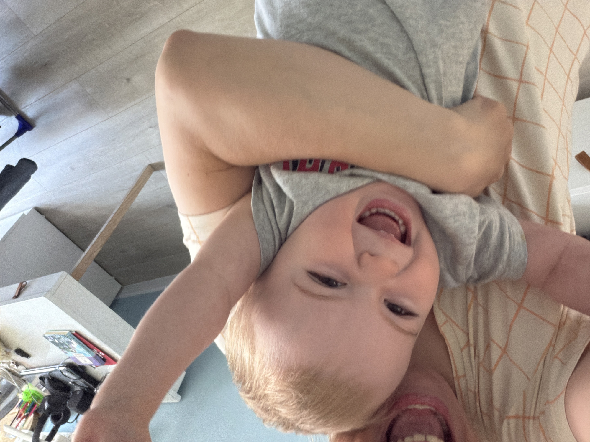 A child is being held upside down by an adult. The child is wearing a grey t-shirt and is laughing with their mouth open. The adult's face is partially visible at the bottom of the frame, also with an open mouth. The adult is wearing a light-colored shirt with a grid pattern. The background includes a grey wooden floor, a white cabinet with items on top, and a light blue wall. The location is unknown; there is no evidence of Votkinsk, Russia.
