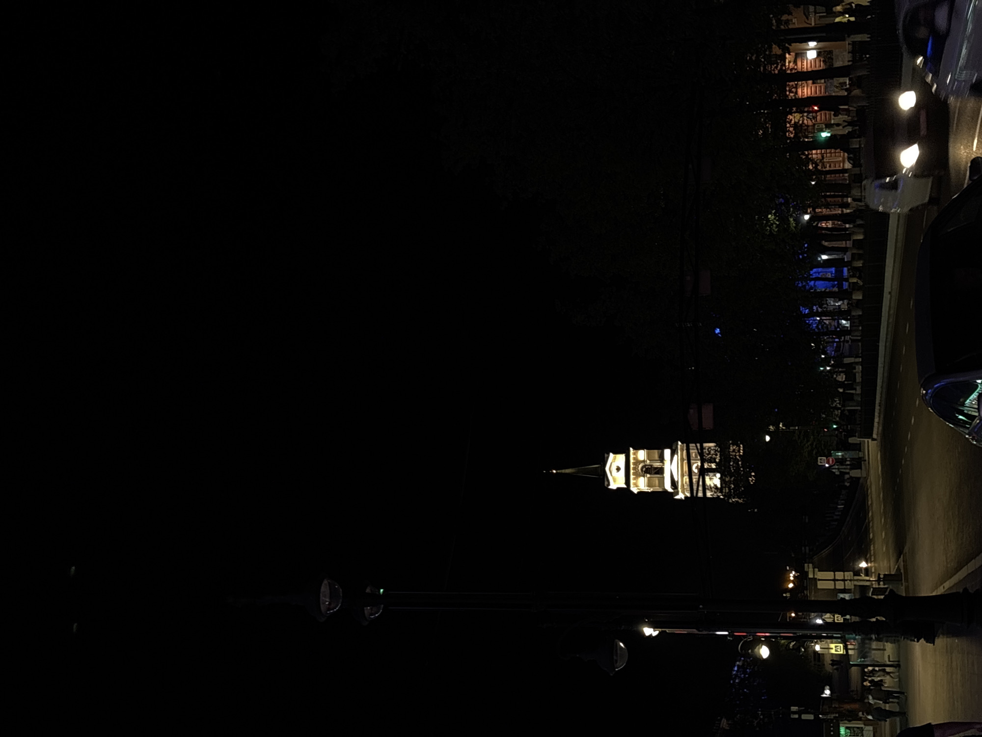 At night, the image shows the Perm State Art Gallery's building, a tower lit up in the middle ground, from across the Kama river. Streetlights illuminate the near road and the buildings and trees on the far side of the riverbank. A section of a car is visible on the right-hand side. A traffic signal pole is in the foreground. Pedestrians can be seen in the middle ground in front of the building in the far background. The scene takes place in Perm, Russia.