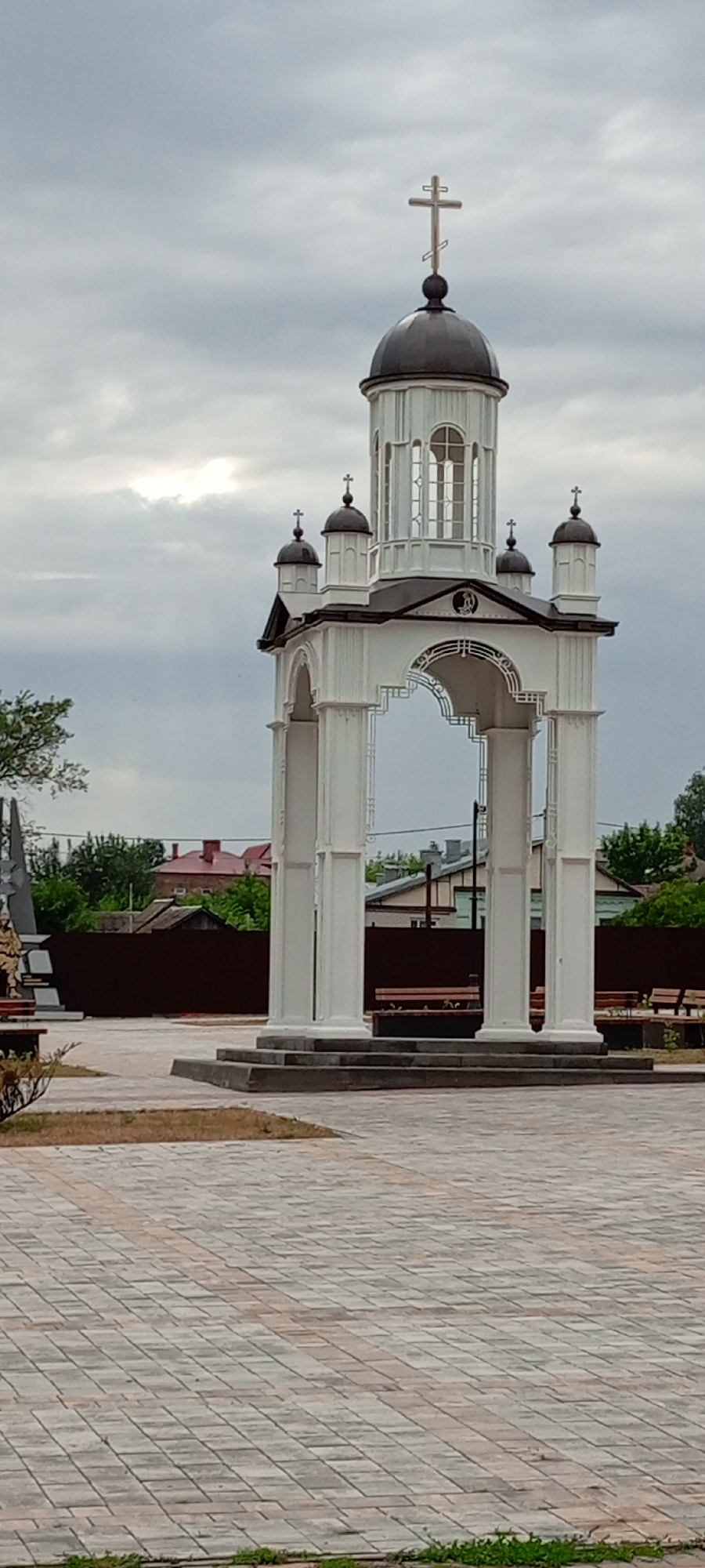 This is a vertical, eye-level, medium shot of an outdoor, open-air pavilion structure topped with an Orthodox cross, suggesting it might be a memorial or a religious site. The structure is white, with four large, fluted columns supporting an arched entryway. Above the arch is a small dome with a cross, and on top of that is a taller structure with a larger, dark dome, also crowned with a prominent Orthodox cross. Smaller domes with crosses are visible on either side of the main dome. The pavilion is set on a paved area made of interlocking bricks, with a darker border. Behind the pavilion is a dark brown fence, beyond which are residential buildings with red roofs and trees. The sky is overcast, with scattered clouds, and the overall lighting is dim. To the left of the pavilion, partially visible, is a monument with a soldier figure and a dark obelisk. Benches are visible behind the pavilion, indicating a public space. The scene appears to be in the city of Kirsanov, Russia, given the architectural style and potential context of the monument.