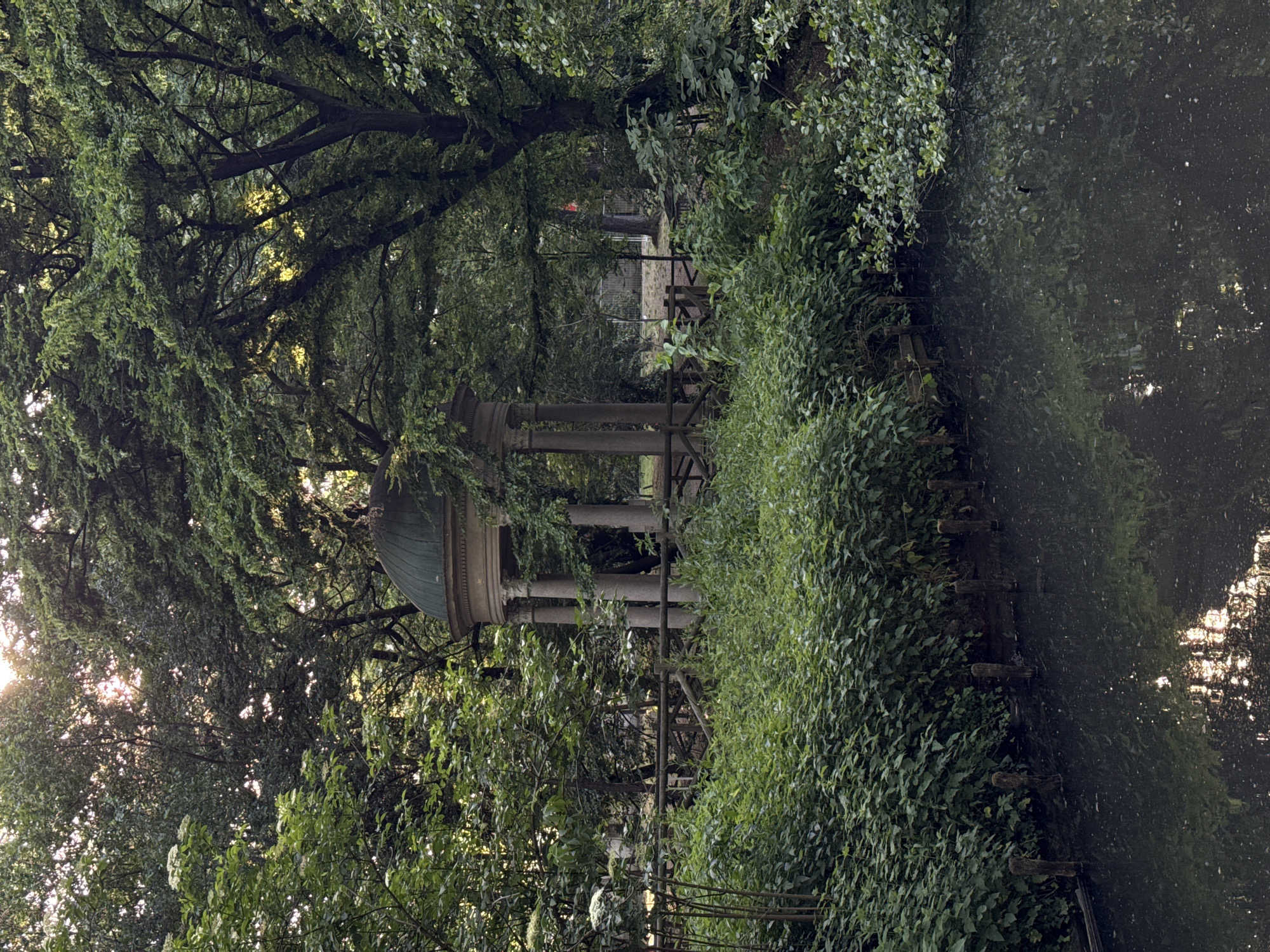 The image captures a tranquil scene in Milan, Italy, likely within the Parco Sempione. A classical pavilion with columns and a domed roof sits amidst lush greenery, framed by trees and overgrown shrubbery. To the right, a body of water, possibly a pond or canal, reflects light from above. The lighting suggests either early morning or late afternoon. The scene is devoid of people, focusing on the architectural element within its natural surroundings. A wooden fence appears to border the pavilion, and glimpses of other structures can be seen in the distance through the trees.