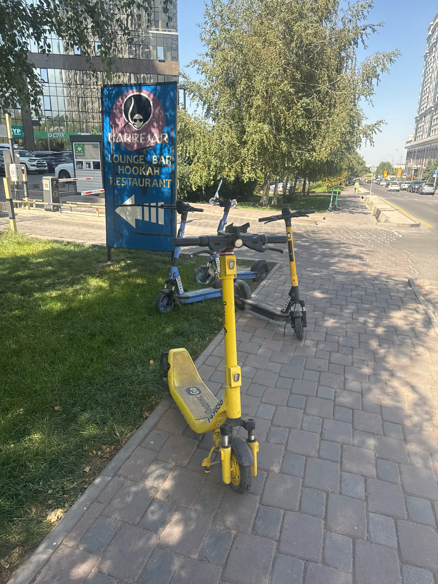 The image shows a street scene in Almaty, Kazakhstan, likely on a bright, sunny day. In the foreground, there are several electric scooters parked on a cobblestone sidewalk. One yellow scooter is particularly prominent, with a license plate reading "Y49856". Behind it, a blue scooter with a license plate "JU9564" is visible. Further back, another blue scooter and a green scooter are parked on a grassy area.
A blue sign with white text indicates "BARREMAR LOUNGE BAR HOOKAH RESTAURANT" and points to the left with a white arrow. Behind the sign, a modern building with a glass facade stands tall, reflecting the sky and trees. The street itself is paved, with traffic visible in the distance, including cars and possibly other public transport. Trees line the sidewalk, casting shadows on the ground, and a few fallen leaves are scattered on the grass. The environment appears to be a developed urban area, with clear blue skies overhead.