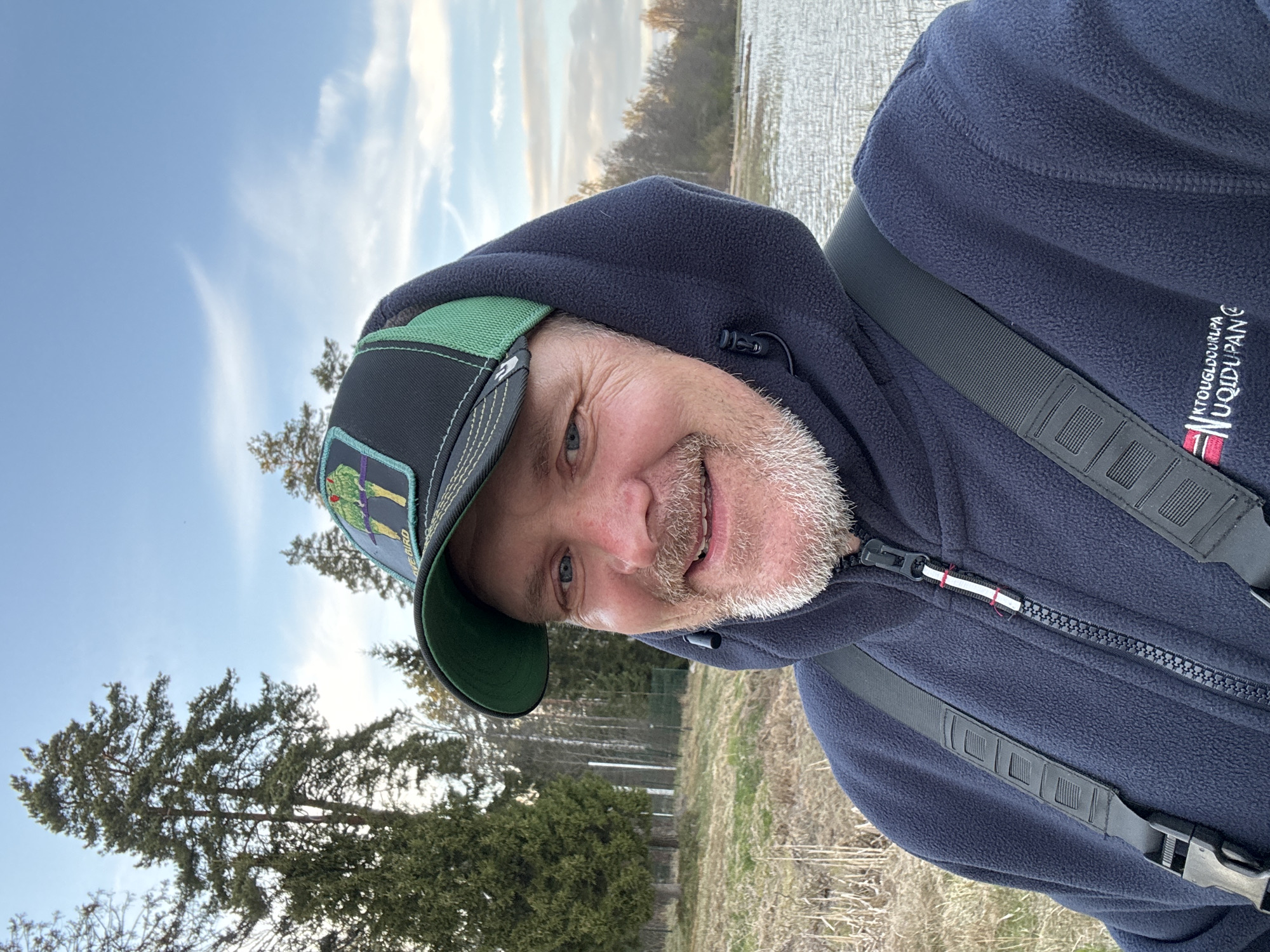 A selfie shows a middle-aged man wearing a dark blue hooded fleece jacket and a baseball cap. The cap is black with a green brim and features a colorful patch. He has a grey beard and light-colored eyes. A black strap crosses his torso, likely from a bag or camera. The background includes trees, a body of water, and a sky with some clouds. The photo seems to have been taken outdoors. It is not possible to establish the location in the city of Aleksandrovka, Russia, from the visible content.