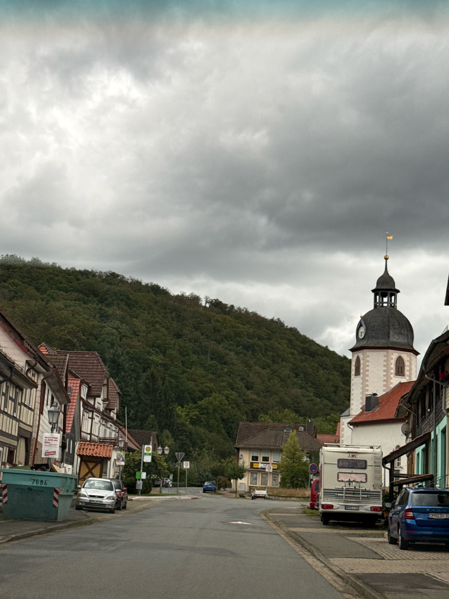 The image depicts a street scene in the town of Bad Sachsa, Germany, under an overcast sky. A steep, densely wooded hill dominates the background, with a prominent church tower featuring a dark, pointed roof and a clock face visible on the right side of the frame. Several half-timbered buildings line the left side of the street, with red-tiled roofs. A large, light-blue dumpster with white diagonal stripes and the number "7604" is positioned on the left edge of the street. A silver sedan is parked near the dumpster, and further down the street, a white recreational vehicle (RV) is parked, with a blue SUV parked behind it. A few streetlights and traffic signs are visible along the street. The overall impression is of a quiet, European town street on a cloudy day.