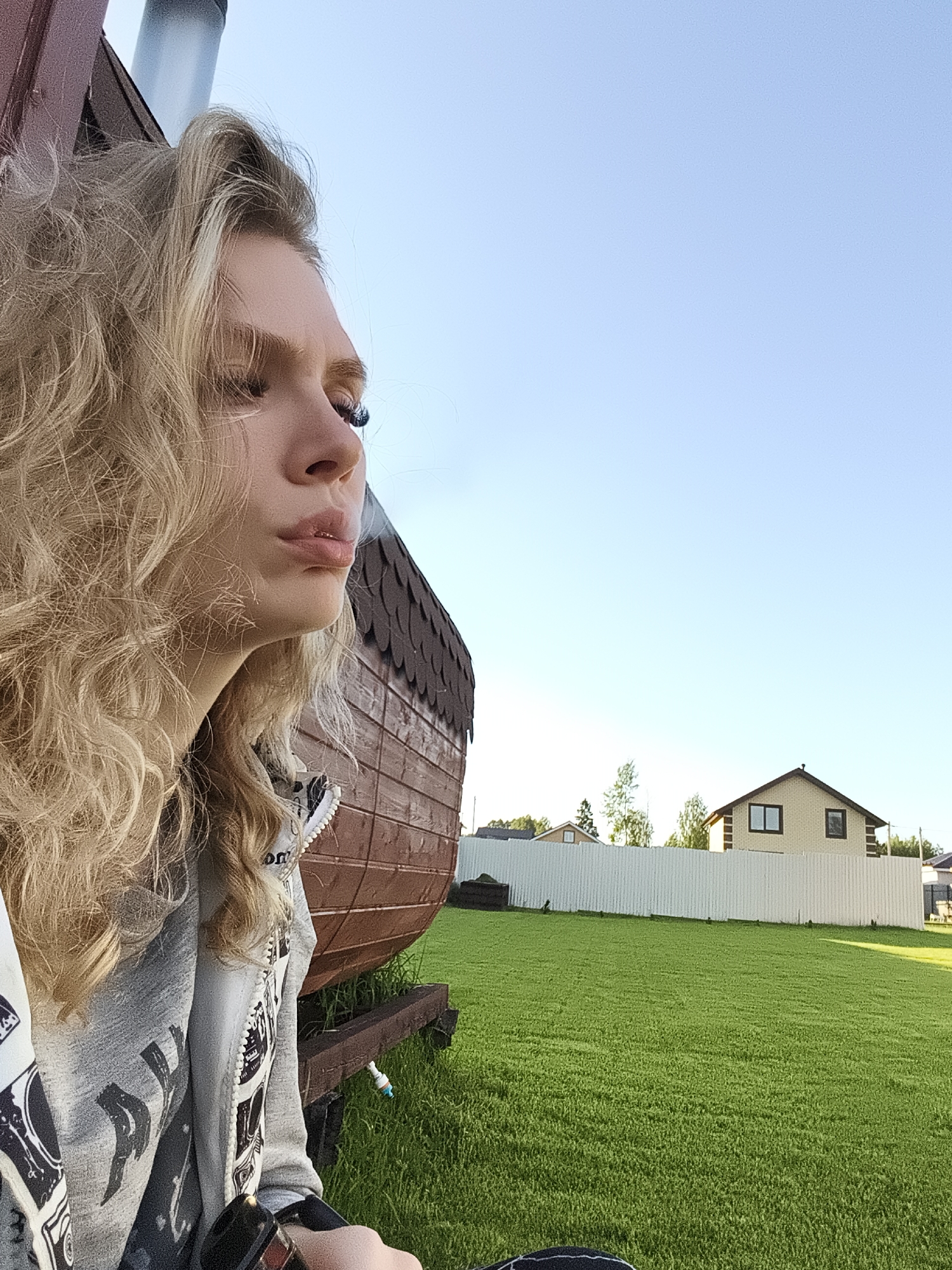This image depicts a young woman outdoors in a rural or suburban setting, likely in Русский Вожой, Russia. She is shown in profile, looking towards the right side of the frame, with long, wavy blonde hair. Her lips are slightly pursed, and a wisp of smoke is visible, indicating she is exhaling from a vape device, which is partially seen in her hand. Her expression appears neutral or pensive.
She is wearing a grey hooded jacket with dark patterns; some text is partially visible on it but is not legible. In the background directly behind her is a rustic wooden structure, possibly a small building or sauna, featuring dark shingled roofing and brown horizontal planks. A small white valve-like component is attached near its base.
The scene opens up to a wide expanse of vibrant green grass in the midground and foreground to the right. A tall, white corrugated fence separates this grassy area from the distant background, where several houses and trees are visible under a clear, bright blue sky. One prominent house is light-colored with dark window frames and a dark roof. The bright light and clear sky suggest it is daytime, possibly morning or late afternoon. The overall impression is one of a relaxed outdoor moment in a calm, open environment.