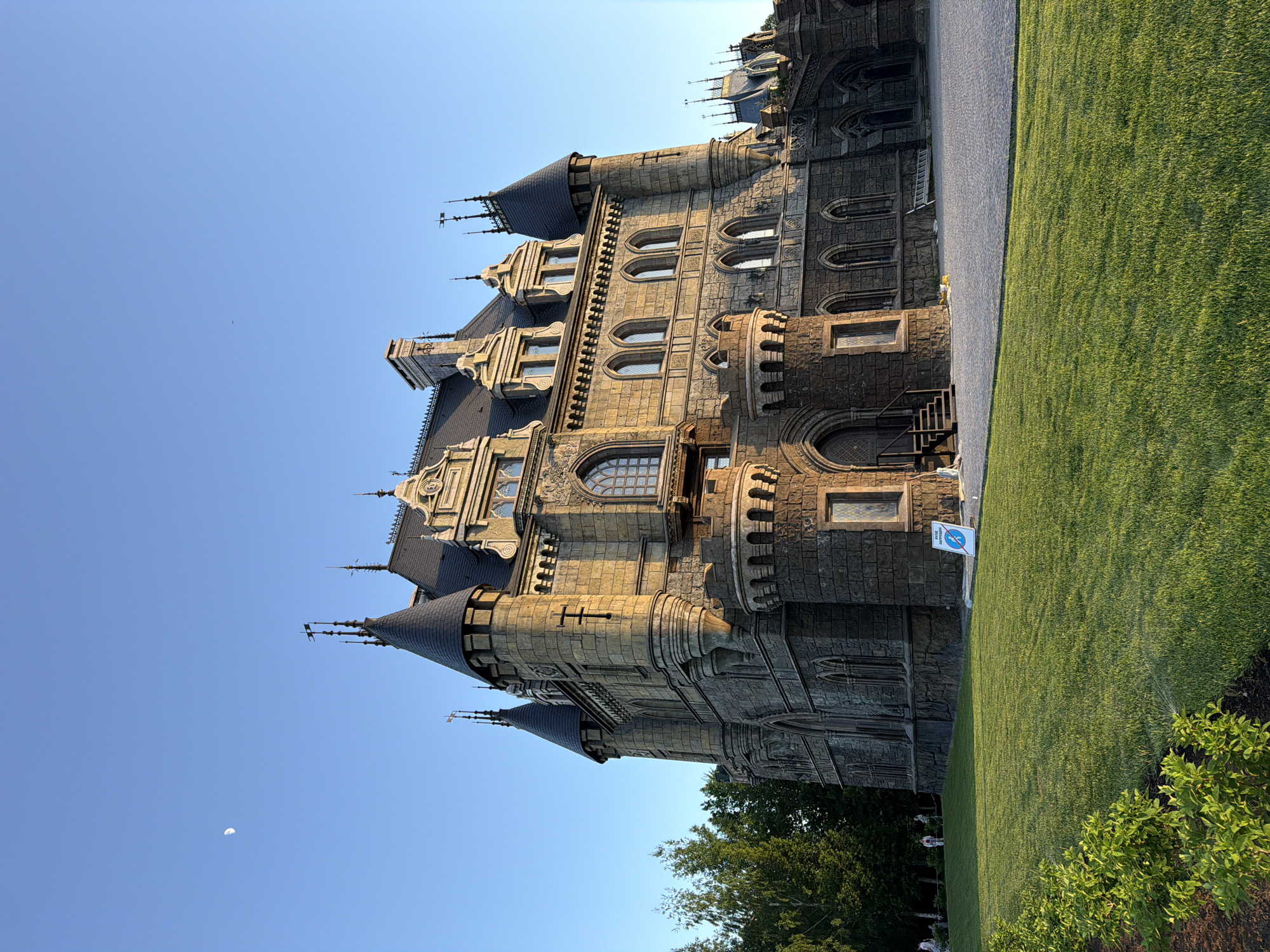 The image shows a large, elaborate castle-like structure, with multiple towers, turrets, and intricate stonework, bathed in warm sunlight. The structure is likely situated in the city of Khryashchevka, Russia. The castle adjoins a lush green lawn, which contrasts with the grey and beige tones of the castle itself. A sign is posted near the lawn. The sky above is clear and blue, and a small crescent moon is visible. Trees are at the base of the image. There are no visible people interacting directly with the structure, though there is someone near the edge of the lawn.