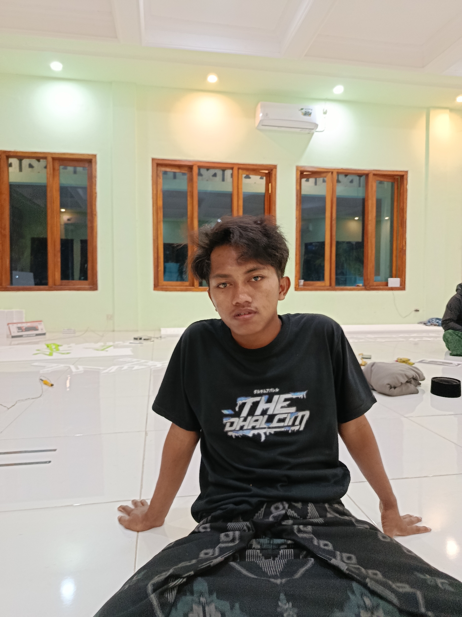 A young man wearing a black t-shirt with a graphic design and a black and grey patterned sarong sits on a white tiled floor in a room with light green walls. Three wooden-framed windows with horizontal panes are visible on the back wall. An air conditioning unit is mounted high on the wall between two of the windows. In the foreground, on the floor to the right of the man, is a black cap. Other objects, including what appears to be a rolled-up blanket or cushion, are present on the floor behind him. The lighting suggests an indoor setting, possibly a community hall or mosque in Ramban Wetan, Indonesia, given the context.
