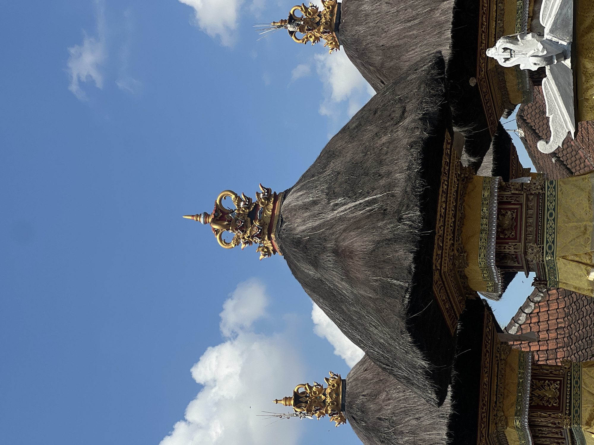 The image captures a close-up view of the ornate thatched roofs of several Balinese structures against a bright blue sky dotted with white clouds. Each roof is topped with intricate golden spires, featuring mythological creatures and decorative elements, characteristic of Balinese temple architecture. The thatch is dark and appears dry, with individual strands clearly visible, giving it a textured, organic appearance. The supporting pillars beneath the roofs are adorned with vibrant yellow and red fabrics, intricate gold patterns, and decorative carvings. In the background, terracotta tiled roofs of other buildings are visible. The scene is static, with no people or animals in immediate focus, highlighting the architectural details and the contrast between the natural materials of the roofs and the detailed golden ornamentation. This architecture is commonly found in religious and cultural sites in North Kuta, Indonesia.