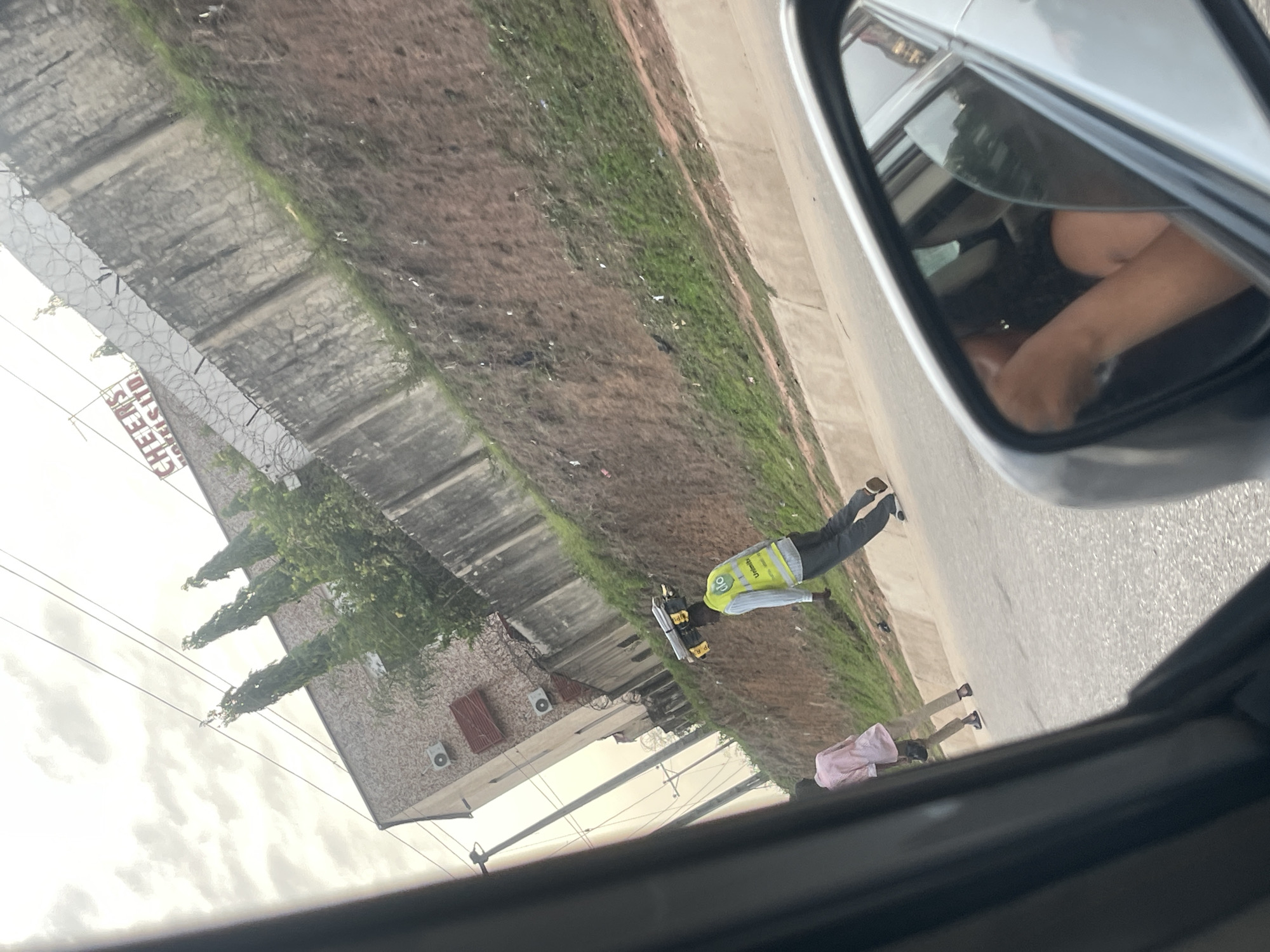 From the perspective of inside a car, the scene shows an exterior street view in Crescent, Nigeria. Along the road, there is a building with a sign that reads "CHEENS". There are two people visible on the road. One is wearing a reflective safety vest, and the other is wearing a pink shirt. A side mirror of the car is also visible.