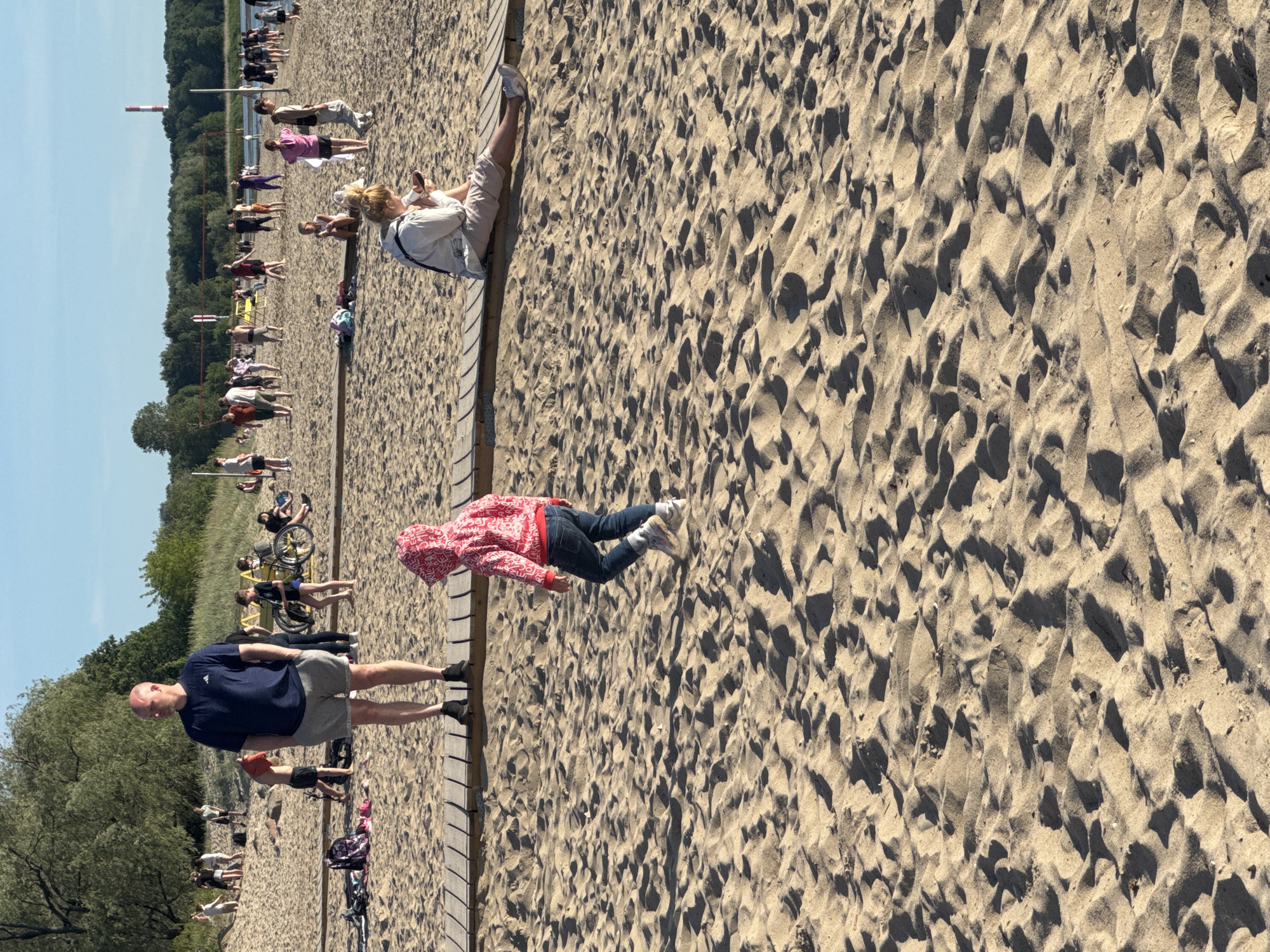 The image depicts a sunny day at Pirita Beach in Tallinn, Estonia. The scene is populated with numerous individuals engaged in various beach activities. In the foreground, a bald man wearing a navy blue t-shirt and grey shorts stands on a wooden walkway, his hands in his pockets, looking to his left. To his right, a young child in a red hooded jacket and blue jeans walks away from the camera. Further back, a woman in light-colored clothing sits on the same wooden walkway, looking down at her phone. The beach itself is sandy, with tire tracks and footprints visible. In the midground, a diverse crowd of people are scattered across the sand. Some are standing and talking, while others are sitting or walking. A group of young people are visible near a volleyball net, suggesting recreational activities. In the background, trees line the horizon, and the sky is a clear blue. The overall atmosphere is one of a relaxed and active summer day at the beach.
