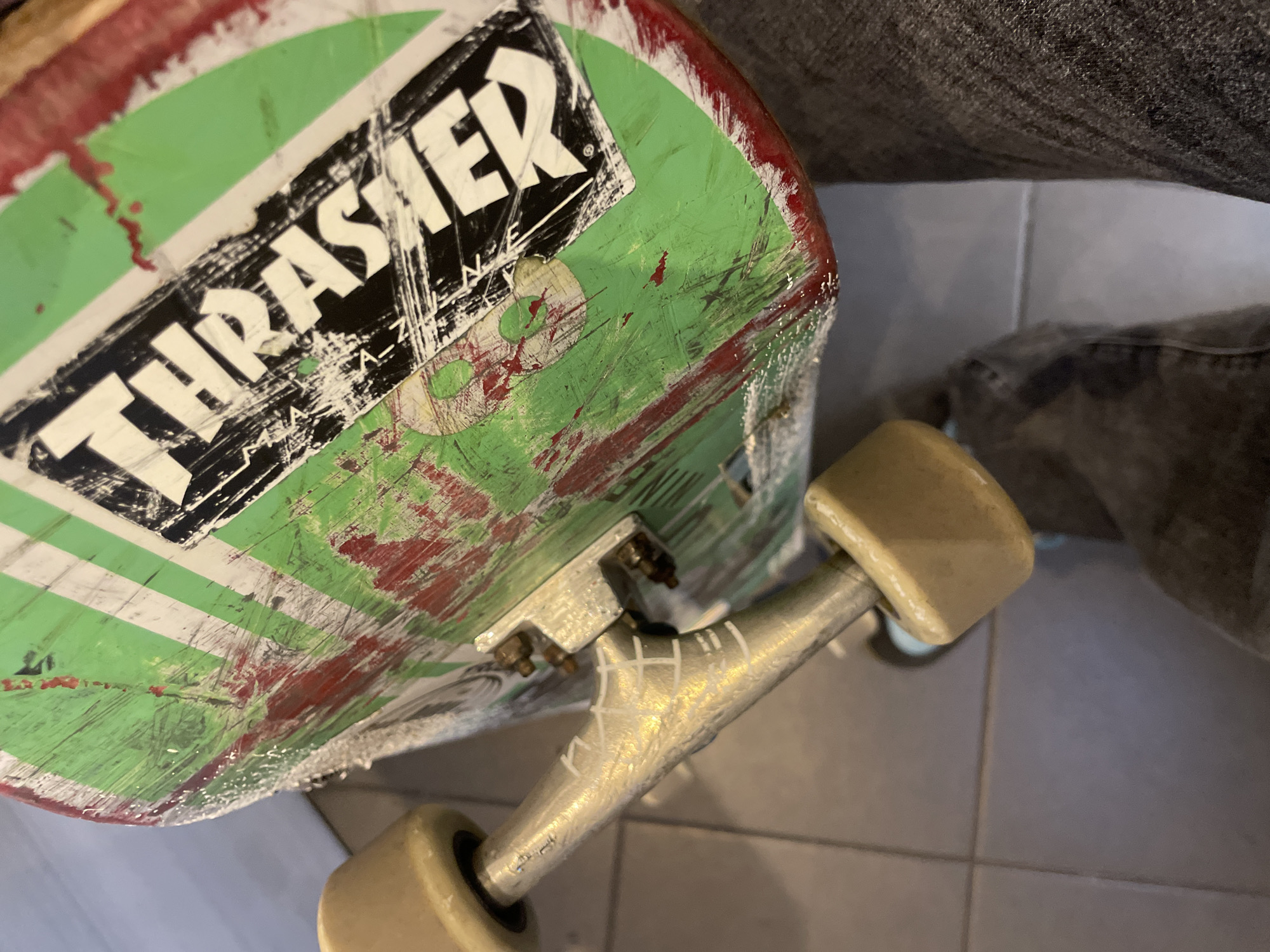 The image shows a worn skateboard with a green, white, and red design, prominently featuring a "THRASHER" sticker on the deck. The board's edges appear scuffed and damaged. It's equipped with silver trucks that have "YKW" etched or written on them in white. Light brown wheels are attached. The background is a tiled floor and a person wearing gray jeans. There is no information in this image to suggest that this is a photo of a location in Barcelona, Spain.