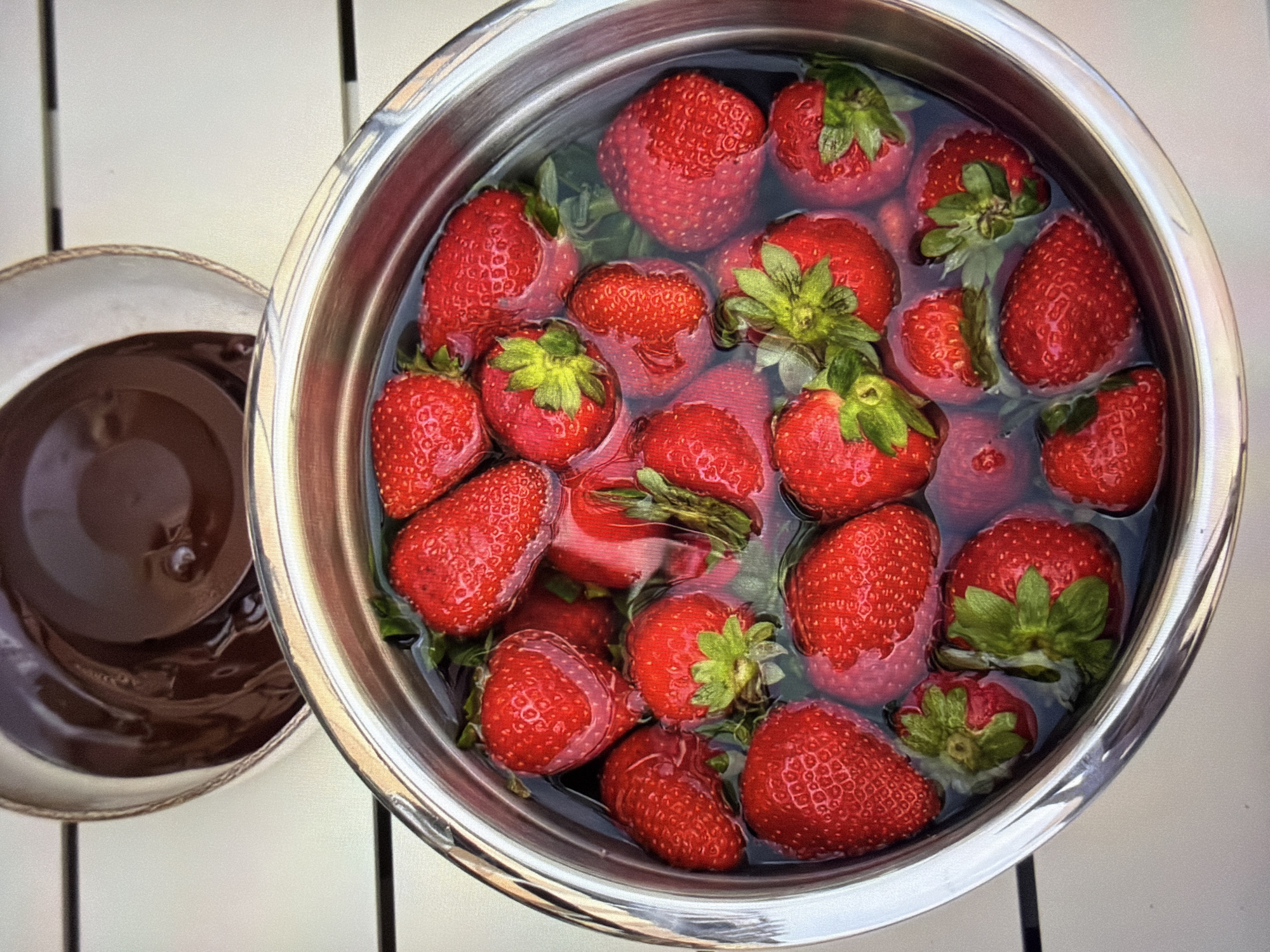 An overhead shot captures two bowls on a bright, possibly outdoor, white slatted surface. The primary subject is a large, silver metallic bowl overflowing with fresh, vibrant red strawberries soaking in clear water. The strawberries, many still with their green caps attached, vary in size and are spread throughout the bowl, some partially submerged, others floating.
To the left of the large bowl, a smaller, dark brown ceramic bowl is partially visible. It contains a thick, glossy dark brown liquid, likely melted chocolate or a similar sauce, with some ripples on its surface.
The lighting is bright and even, indicating daylight, possibly outdoors on a patio or garden table. The white surface underneath the bowls appears to be made of planks or slats, creating subtle linear shadows. The overall scene suggests the preparation of a dessert, such as chocolate-covered strawberries, with the strawberries being washed before dipping. No people or visible text are present in the image. The location is Ros Na Rí, Ireland.