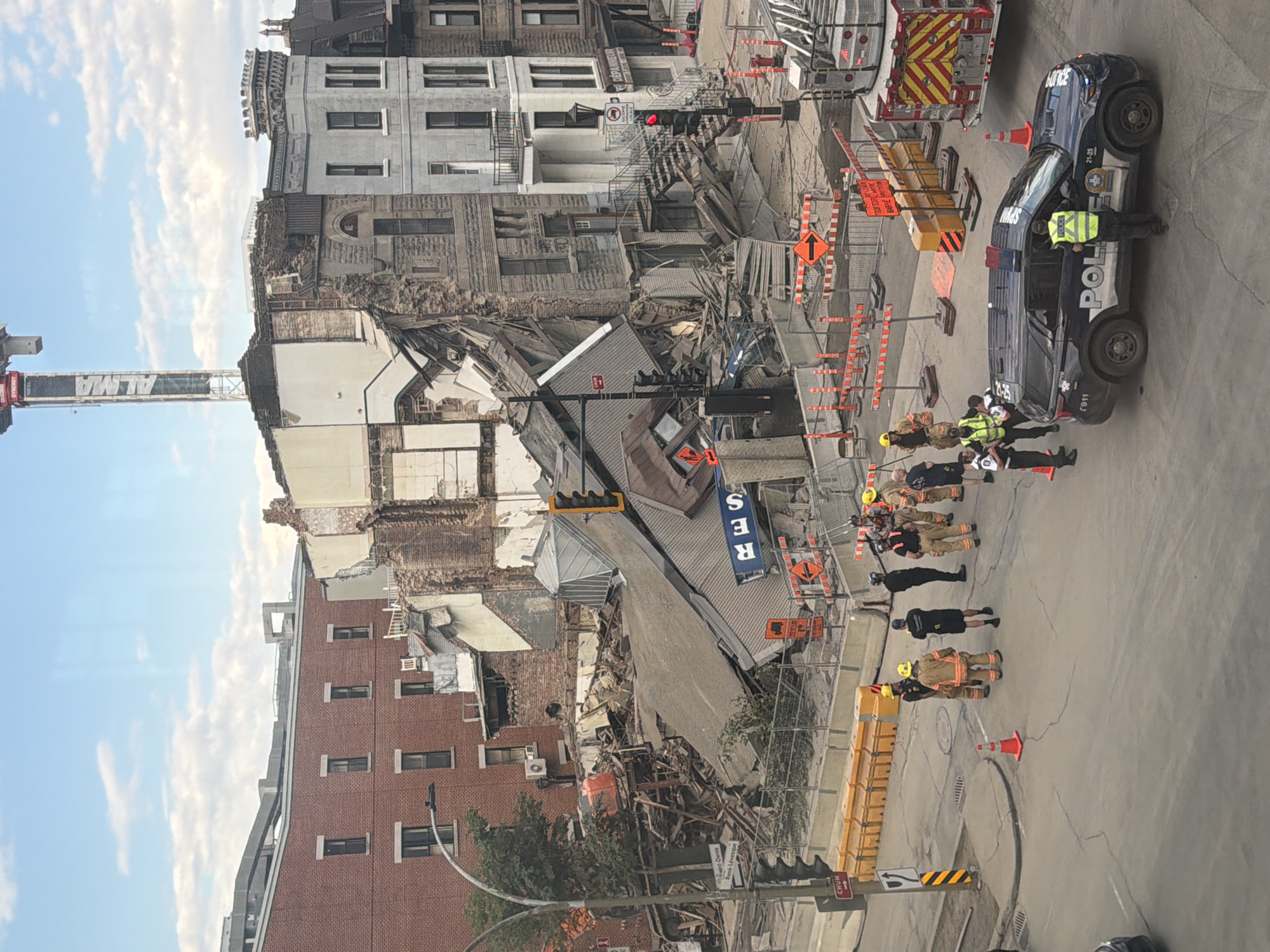 This media file captures a scene of destruction and emergency response in Montreal, Canada. The primary focus is a partially collapsed building, with debris scattered across the street and surrounding area. Several emergency personnel, including firefighters and police officers, are visible attending to the scene. A police car with "POLICE" and "911" clearly marked is present, along with a fire truck in the background.
The setting appears to be a city street, with buildings lining the road. Construction barriers and traffic cones indicate that a work zone or a cordoned-off area is in effect. The sky is blue with some clouds, suggesting it is daytime. The overall atmosphere conveyed is one of serious incident and ongoing recovery efforts. Visible text includes "RES", "DÉPANNEUR", "POLICE", "911", and street names like "CHUM" and "René Lévesque". The presence of the "CHUM" sign could indicate proximity to the Centre hospitalier de l'Université de Montréal. The image is oriented vertically.