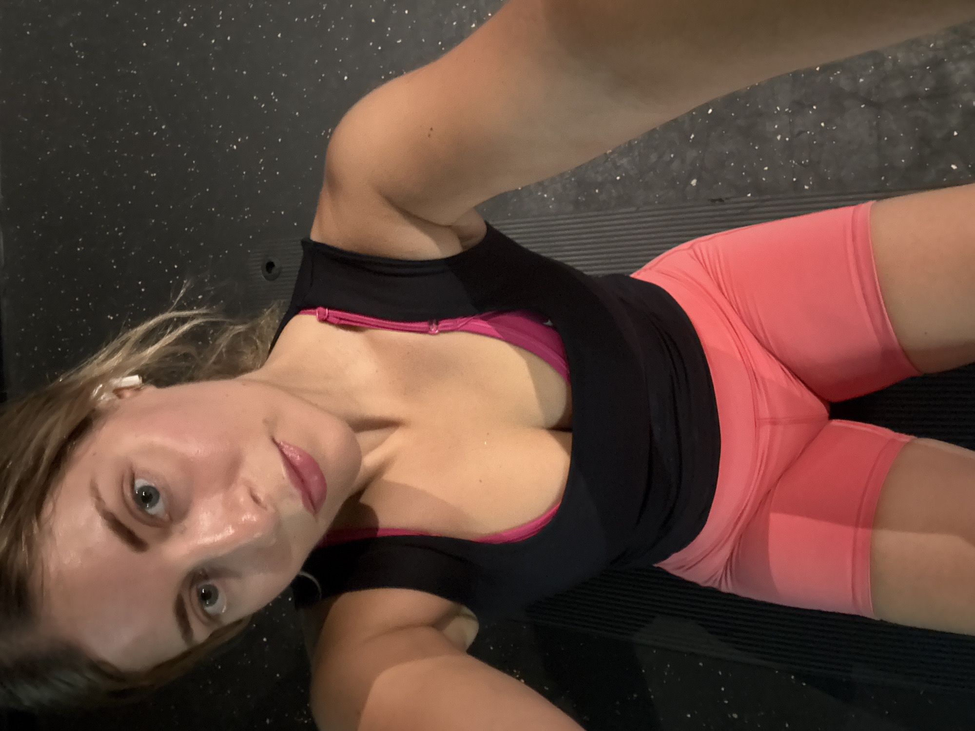A high-angle, close-up shot shows a woman lying on a dark gray textured surface, likely a gym floor. She is wearing a black tank top over a pink sports bra and bright pink athletic shorts. Her face is illuminated, and she appears to be looking at the camera. She has blonde hair and is wearing white earbuds. The background is dark and out of focus, with some speckled white elements visible on the surface. There is no discernible location in Paris, France.