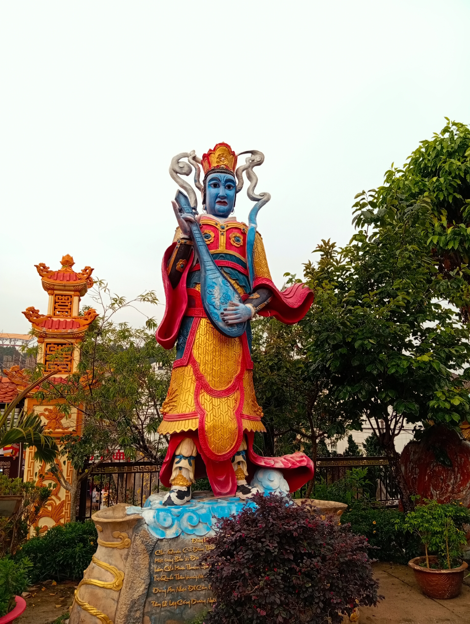 An outdoor, eye-level shot captures a vibrantly colored statue of a deity or guardian figure within a garden setting at Cam Ly, Đà Lạt, Vietnam. The central subject is a standing statue featuring blue skin, red lips, and a serious expression, adorned in an elaborate red and golden robe with intricate patterns and a matching crown. The figure holds a blue stringed instrument, resembling a pipa or lute, decorated with carved motifs.
The statue rests on a stone base embellished with blue and white cloud-like elements. Gold Vietnamese text is inscribed on the base, including phrases such as "Dang," "Chỉ Quản Côn Đông T," "Một trong Bến Vị Độ," and "Dùng Âm Nhạc Độ Càng tỉ" (using music), which hint at the figure's identity or purpose, possibly one of the Four Heavenly Kings associated with music.
In the mid-ground to the left, an ornate, multi-tiered pagoda-like structure with red and orange roofs and intricate details stands amidst green foliage. Lush green trees and bushes frame the statue on the right and behind, while darker red-leafed bushes and potted plants occupy the immediate foreground. A dark metal fence with a geometric pattern is visible behind the statue's base. The sky is overcast and uniformly light, suggesting a diffused light scenario. The scene presents a cultural or religious site with a serene atmosphere.
