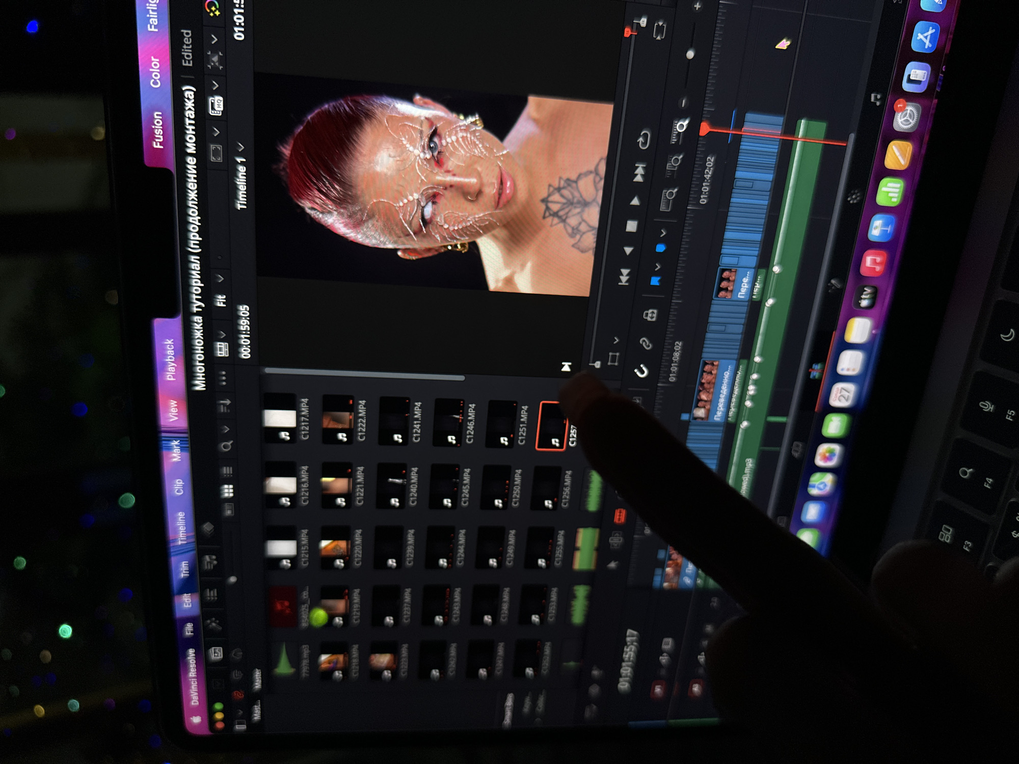 The image shows a laptop screen displaying DaVinci Resolve software, likely being used for video editing. The software interface is visible, including a timeline, a video preview window, and a media pool with numerous video clips. The video preview shows a woman with striking makeup and body art. The laptop is resting on a table with colorful, blurred lights in the background. A finger is pointing to one of the video clips in the media pool. The location is not discernible from the given image content and cannot be determined to be in North Kuta, Indonesia.