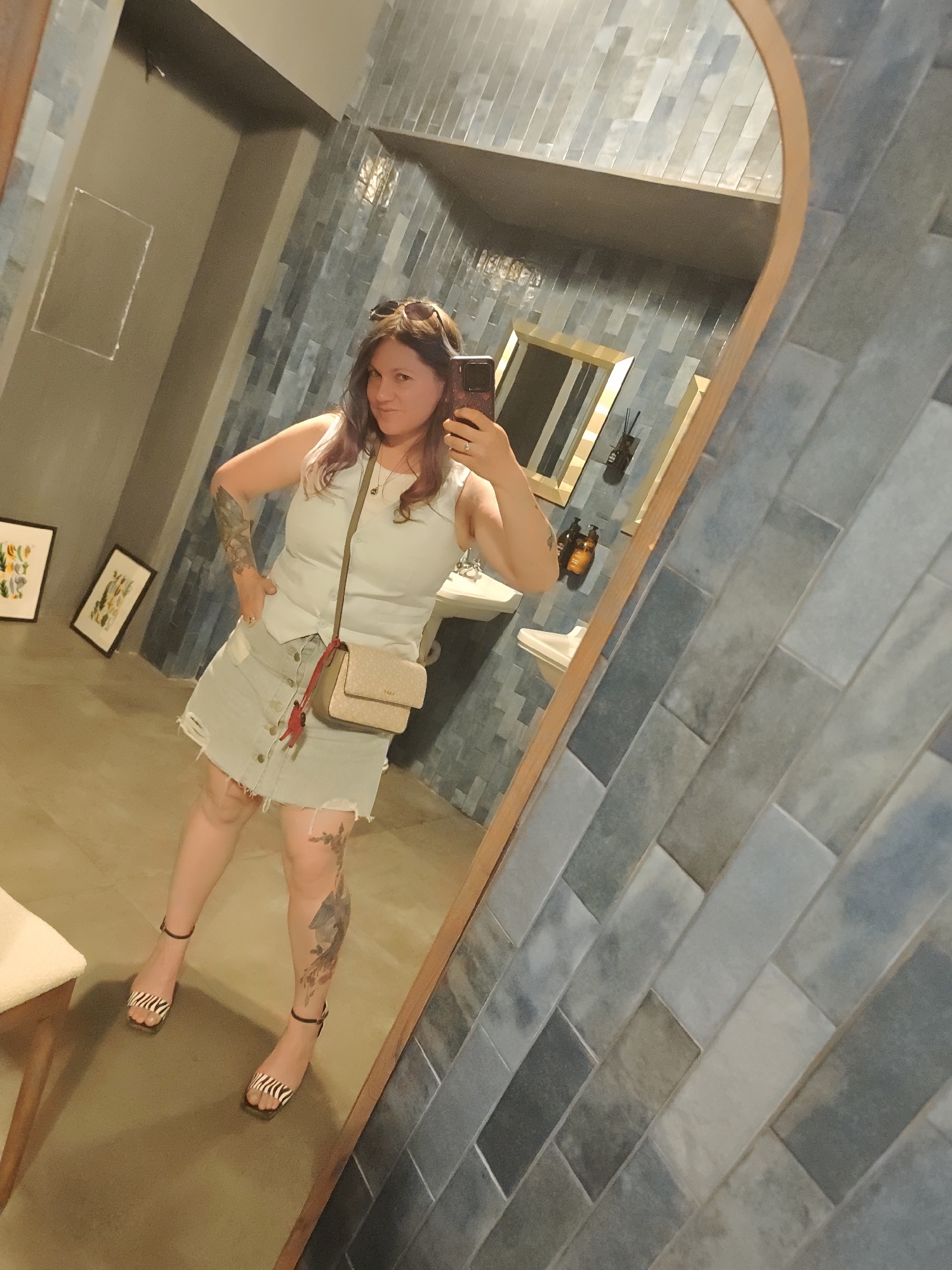 A woman is taking a mirror selfie in what appears to be a restroom. She is wearing a light-colored tank top, a distressed denim skirt, and zebra-print heeled sandals. A small crossbody bag with a light-colored strap and a beige flap with a logo is slung across her body. She has tattoos on her arms and legs. The restroom features blue, grey, and white rectangular tiles on the wall and a large, oval mirror with a wooden frame. In the background, a sink and a small shelf with bottles are visible. Two framed pictures hang on the wall to the left. The floor is a neutral color. The scene does not provide any specific indicators of its location within Moscow, Russia.