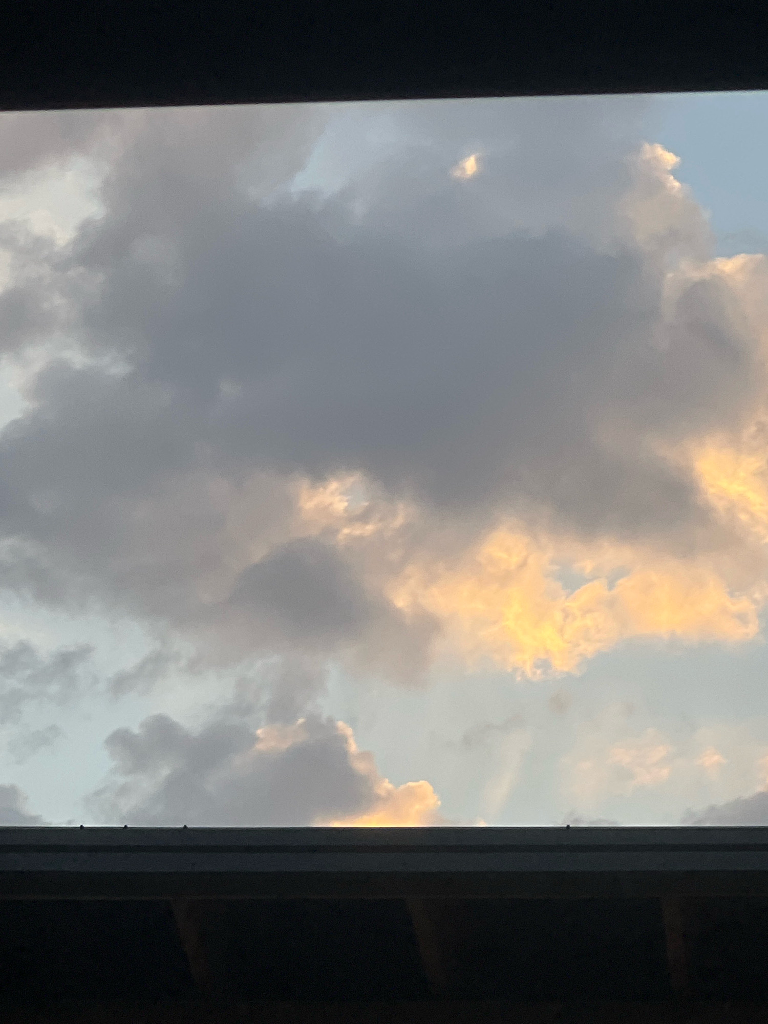 The image captures a sky filled with clouds, some appearing gray and overcast, while others are illuminated by a warm, golden light, suggesting either sunrise or sunset. The scene is framed by the dark silhouette of a structure along the bottom and top edges of the frame, possibly a roofline or awning. The location is stated as Unknown, Unknown. There are no people, animals, or specific landmarks visible in the image, only the interplay of light and clouds against a dark, man-made foreground.