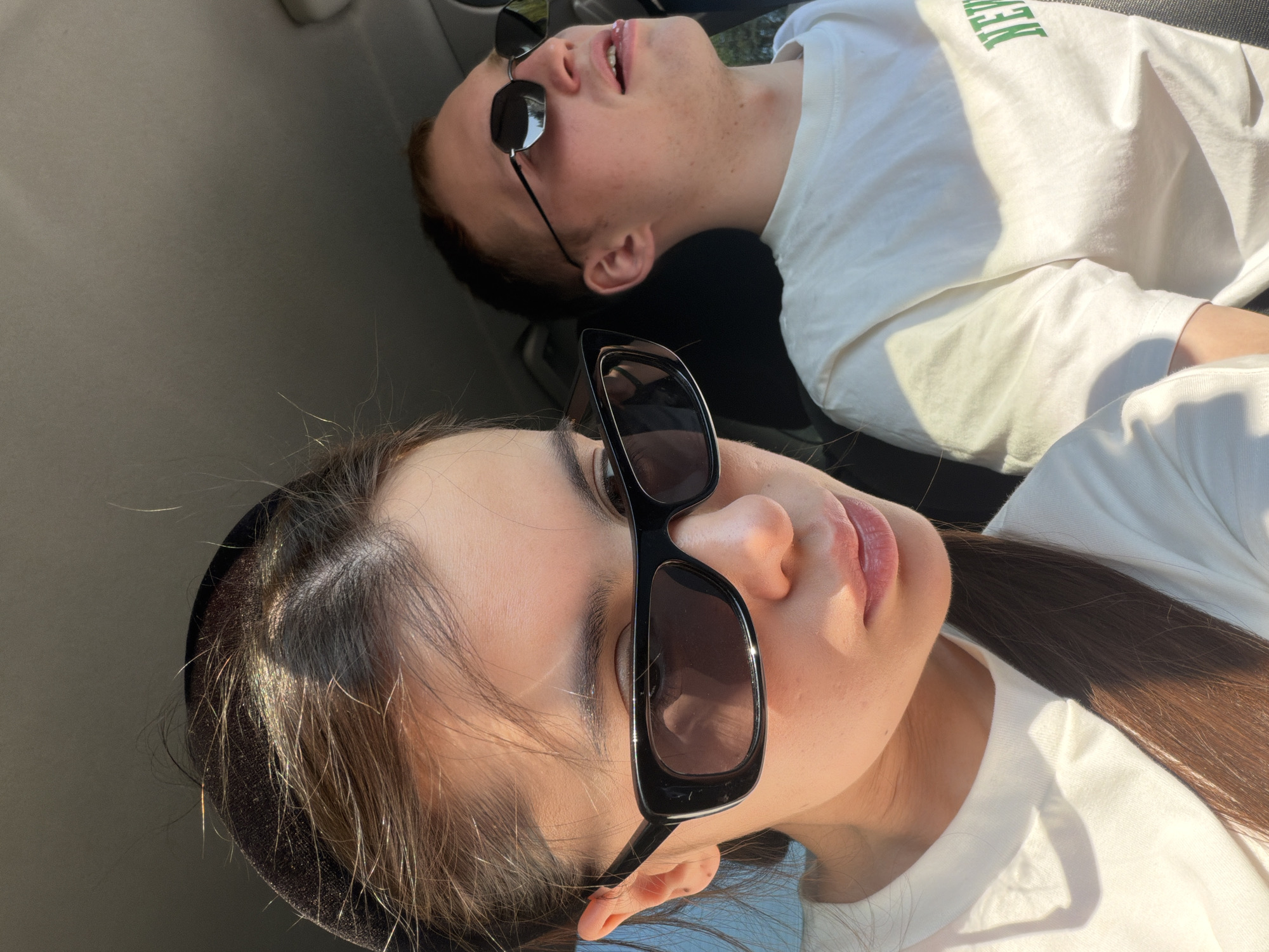 Two people are reclining in a car. A woman with dark hair and black sunglasses is in the foreground, while a man with short hair and round sunglasses is in the background. Both are wearing white shirts. The car's interior is visible, including the headliner. The location is not discernible from the image.