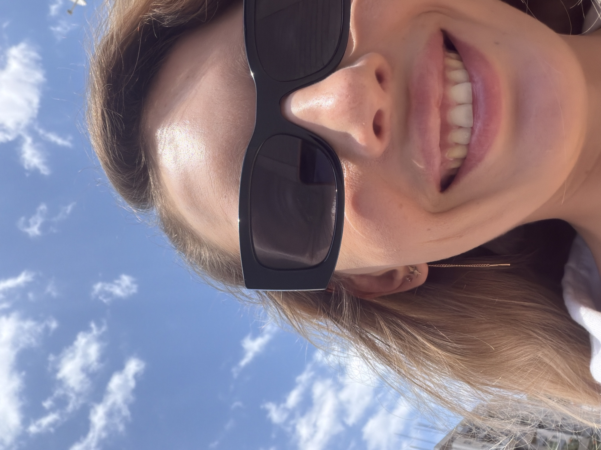 The image is a close-up, eye-level selfie of a young woman with blonde hair, wearing large, black, rectangular sunglasses. She is smiling, showing her teeth, and her mouth is slightly parted. Her left ear is visible, adorned with a small star-shaped earring with a dangling chain. The background is a bright blue sky with scattered white clouds. The upper portion of buildings, likely in a city, is visible on the left side of the frame. The lighting suggests it is daytime, possibly outdoors. The photographer's location is not determinable from the image itself.