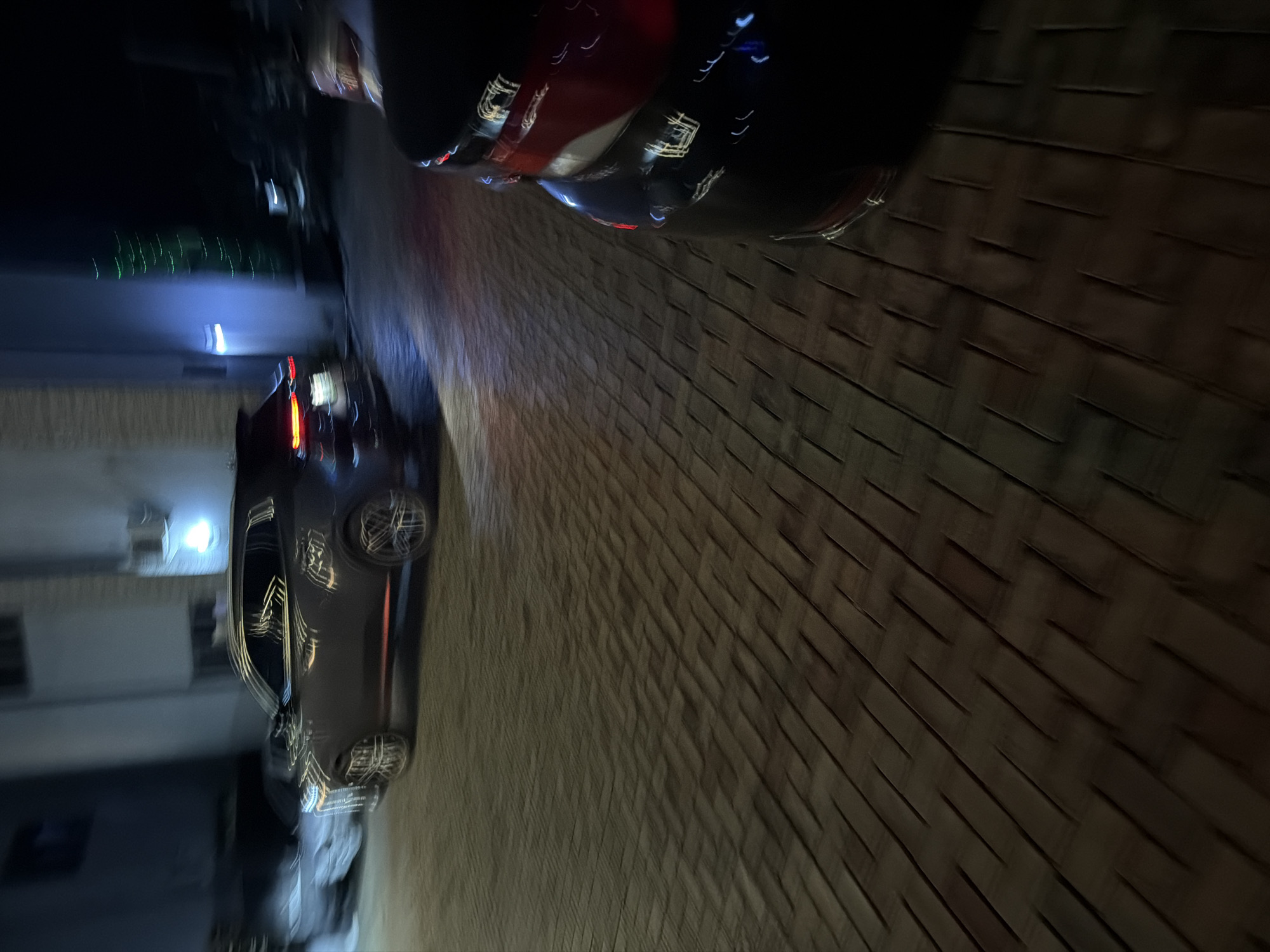 The image depicts a night scene in a residential or commercial area in Bukuru, Nigeria. The foreground is dominated by a dark-colored car, with its rear visible and reflecting ambient lights. To the left, a dark SUV is parked or in motion, its headlights and side profile illuminated. The ground is paved with brickwork, showing a pattern of staggered rectangular bricks. In the background, a building with illuminated windows and a blue light fixture is visible. The overall impression is that of a street or driveway at night, with cars and architectural elements illuminated by artificial light sources. The motion blur suggests movement within the scene.