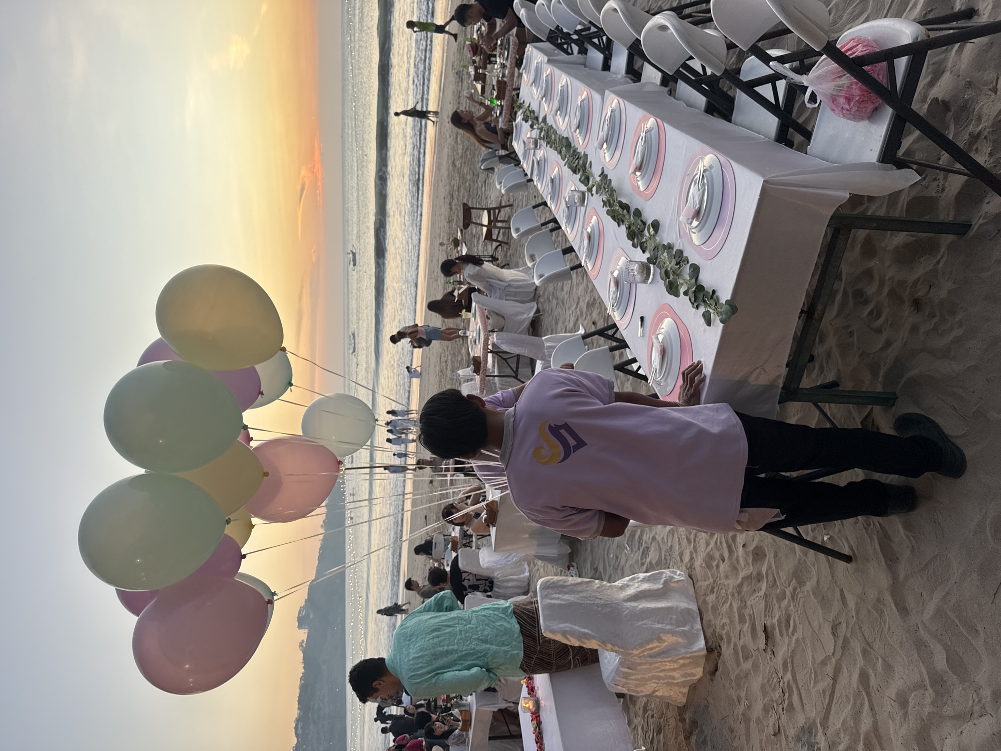 A group of people are gathered on a beach in Kuta Selatan, Indonesia, for an outdoor event at sunset. A long table is set with white tablecloths, plates, and cutlery, decorated with a garland of greenery. White chairs are arranged around the table. In the foreground, a man in a light purple shirt with a logo on the back faces away from the camera, and another man in a teal shirt stands near a table covered in white fabric. In the background, the ocean meets the sky in a colorful sunset, with a few small boats visible on the water. More tables and chairs are set up on the beach, and a cluster of pastel-colored balloons floats above the scene. Several other individuals are scattered throughout the background, some dining at tables and others walking along the shore. The sandy beach extends to the water's edge.
