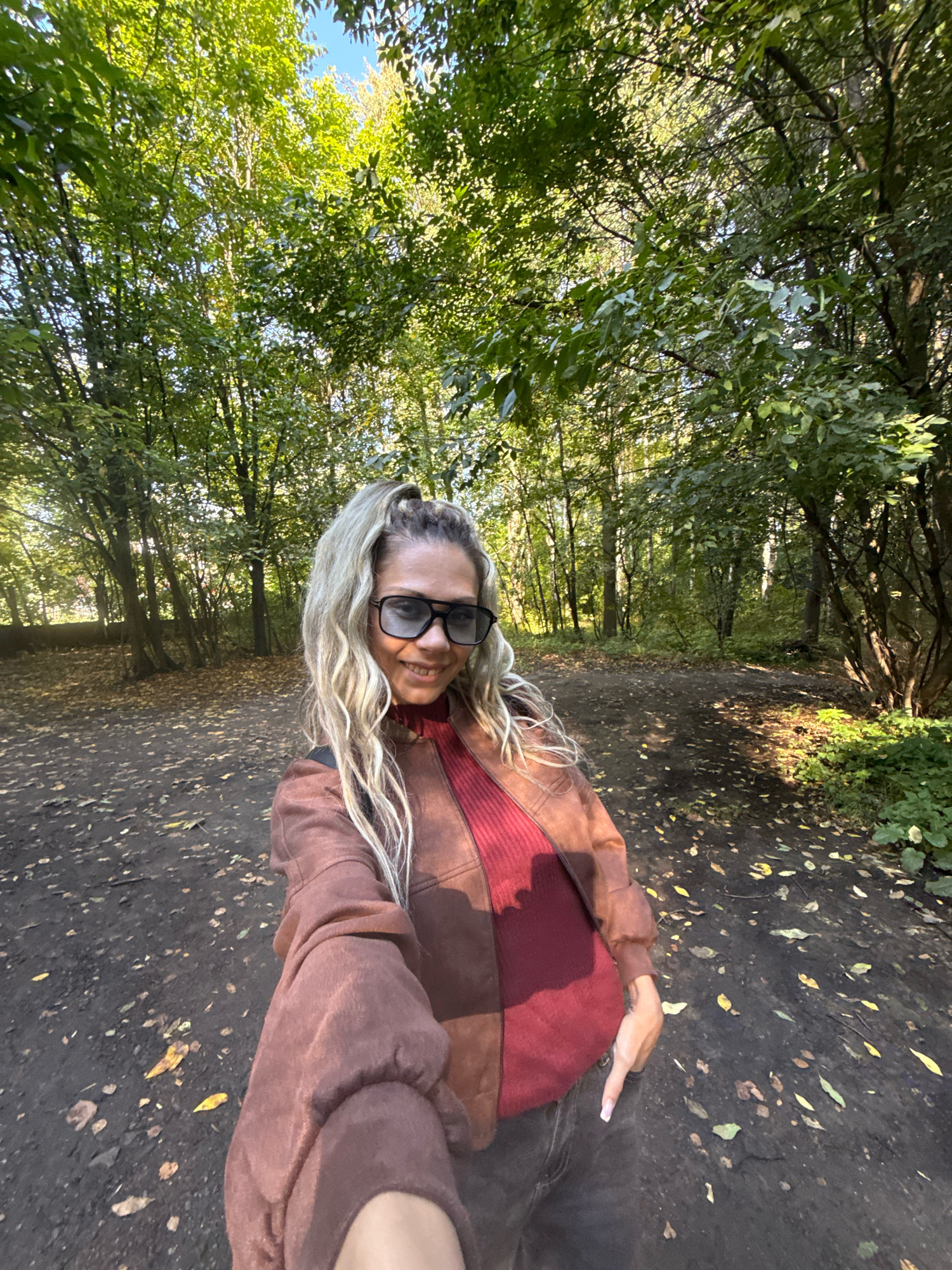 A woman with blonde hair pulled into a ponytail and wearing large dark sunglasses, a rust-colored jacket over a red turtleneck, and dark pants stands in a dirt path in a wooded area. Fallen leaves are scattered across the ground. The trees are green and lush, with sunlight filtering through the canopy. The background shows more trees and a hint of blue sky. The image is a selfie, taken from the perspective of the woman holding the camera. There is no identifiable context relating to Rusanovo, Russia.