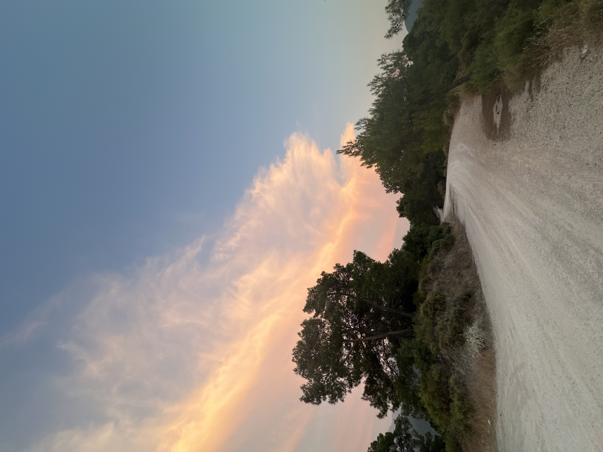 The image depicts a dirt road curving through a landscape of trees and scrub under a sky with soft, peach-colored clouds. The road appears to be in a rural or mountainous area, possibly on the outskirts of Fethiye, Turkey. The lighting suggests it is either early morning or late afternoon, with the sun casting a warm glow on the clouds. There are no people or animals visible in the scene. The environment is characterized by lush green vegetation and dry, earthy terrain. The road itself is made of gravel and dirt, indicating it may be a less-trafficked route. The overall impression is one of natural beauty and tranquility.