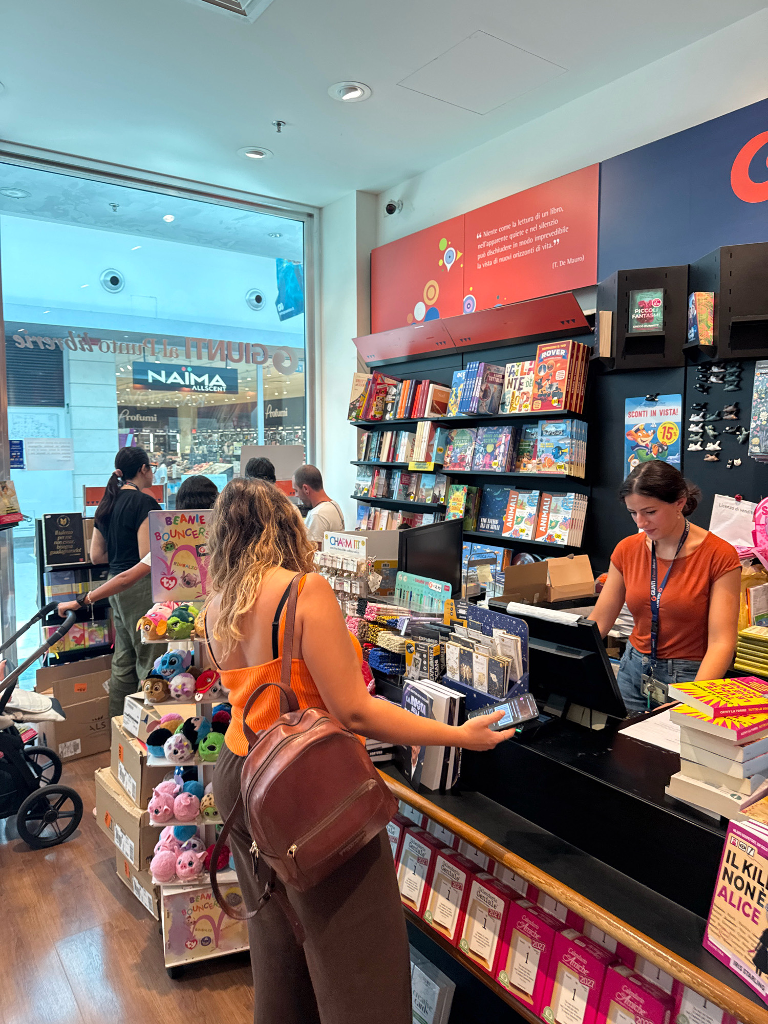 The image depicts the interior of a bookstore, likely in the city of Carini, Italy, indicated by the text visible on signs. Several people are present. In the foreground, a woman wearing an orange top and a brown backpack is interacting with a cashier at the checkout counter. She is holding a smartphone, possibly making a payment. The cashier, also a woman, is wearing an orange shirt and blue jeans. Behind them, shelves are filled with books and various merchandise, including plush toys. Through a large window on the left, an outdoor shopping area with other stores is visible. The overall environment suggests a busy retail setting.