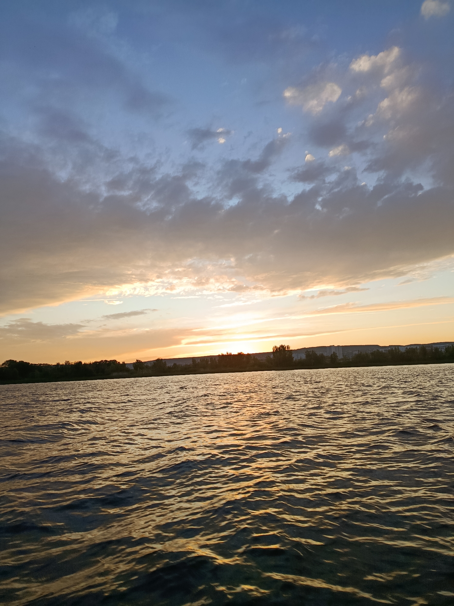 This is a horizontal, eye-level, full shot of a body of water with a sunset sky in the background. The water is choppy and reflects the golden light of the setting sun. The sky is a mix of blue and orange, with clouds scattered throughout. In the distance, a treeline is visible with buildings and structures interspersed among the trees. The scene appears to be a natural body of water such as a river or lake, likely on the outskirts of a city. Given the description of Saratov, Russia, this could potentially be the Volga River. There are no people or animals visible in the frame, and no discernible actions or interactions.
