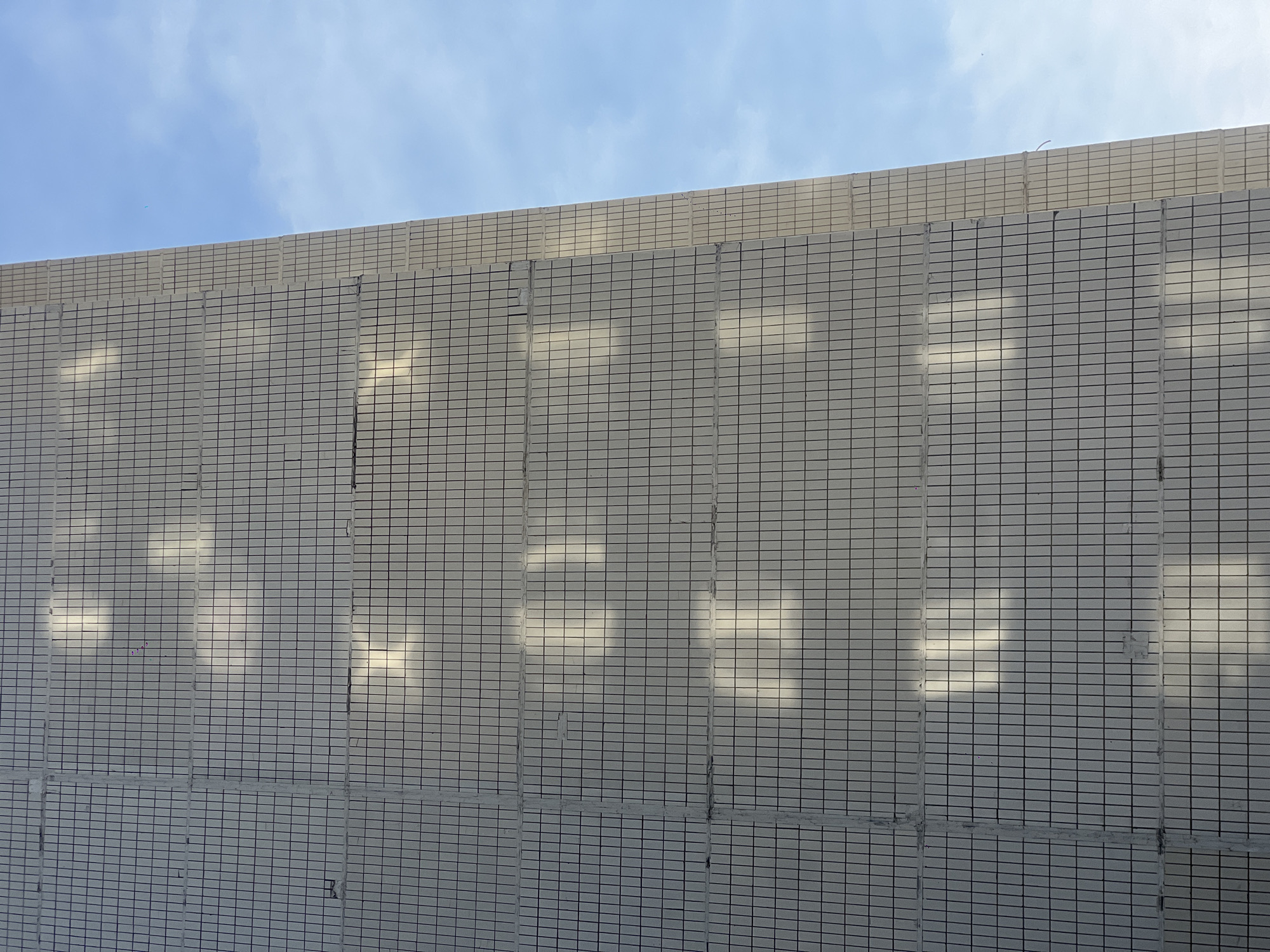 The image shows a section of a building exterior made of light-colored rectangular tiles with dark grout lines. Sunlight creates a pattern of brighter areas across the tiled surface, seemingly projecting the shapes of nearby objects or structures. The sky is visible in the upper left corner, revealing a light blue color with some clouds. No people or other specific objects are clearly visible. There is no indication of the location being Moscow, Russia. The scene appears to be a static, architectural detail captured in daylight.