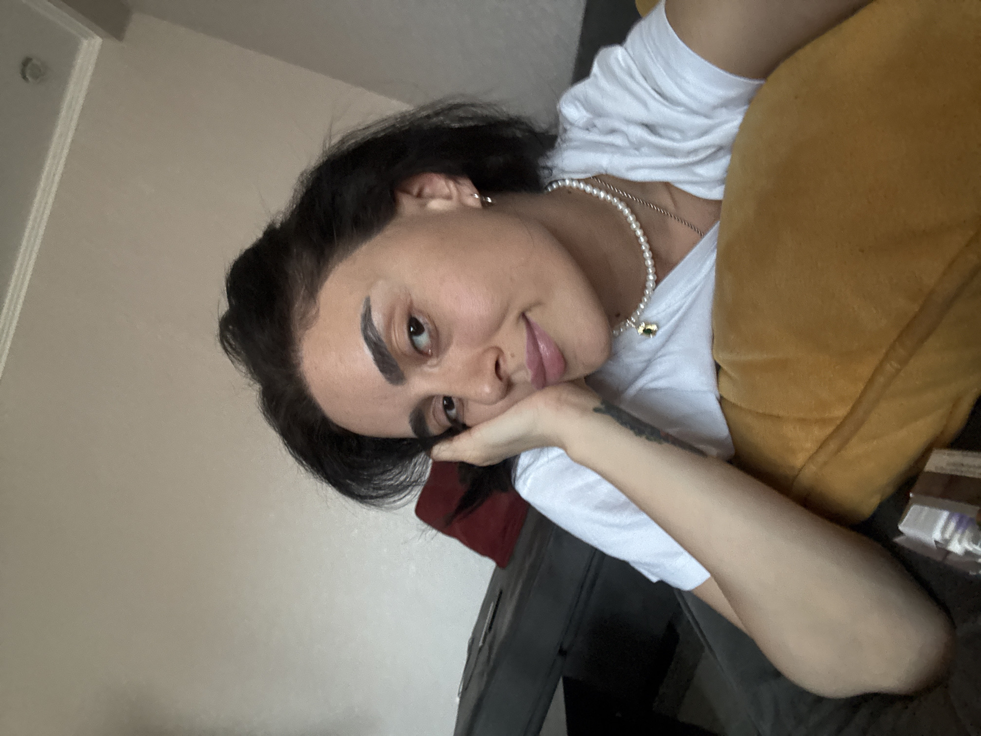 A medium shot captures a woman with dark hair and light skin from the shoulders up. She is resting her head on her left hand, with her right arm bent and resting on a yellow pillow. She wears a white t-shirt, a pearl necklace, and a small pendant. Her eyes are looking forward, and her lips are slightly parted. A blurred background shows a grey sofa and a wall. There is no indication of Tbilisi, Georgia.