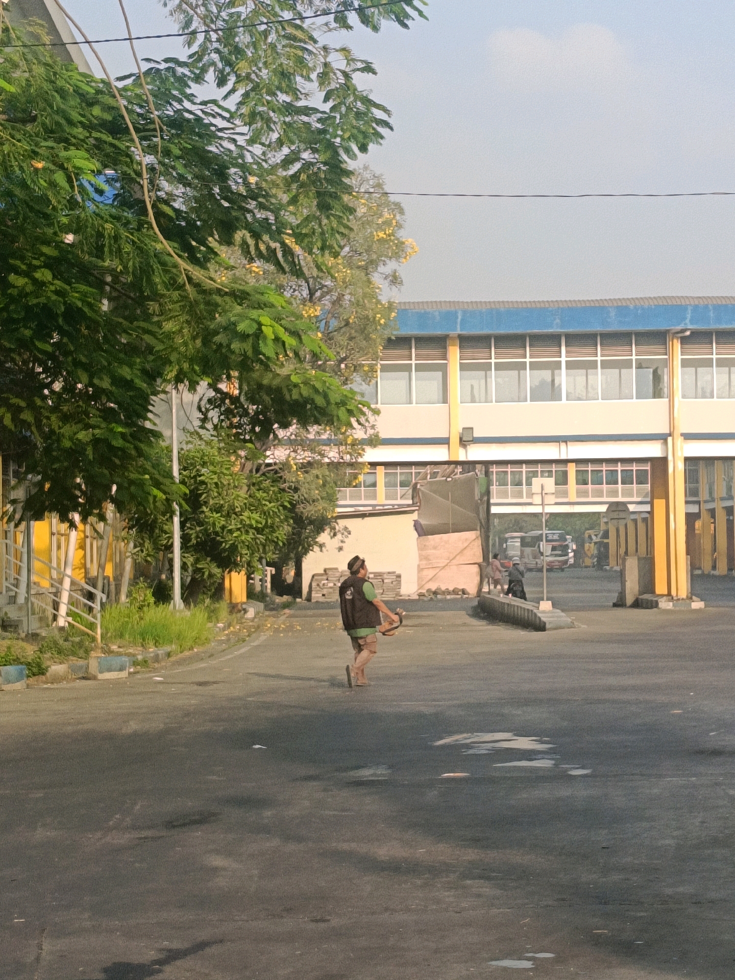 A man wearing a dark vest over a green shirt and light brown shorts walks on an asphalt road in Kedungrejo, Indonesia. He is carrying a long object, possibly a broom or a tool, horizontally across his body. The road is lined with grass and concrete barriers, some of which are painted blue and white. In the background, a modern building with a blue roof and white window frames stands. The building has multiple stories and an open entryway where several buses are visible. Trees with green leaves and some yellow blossoms are scattered throughout the scene, and power lines run across the sky, which is a pale blue with some clouds. The ground appears damp in places, suggesting recent rain or watering.