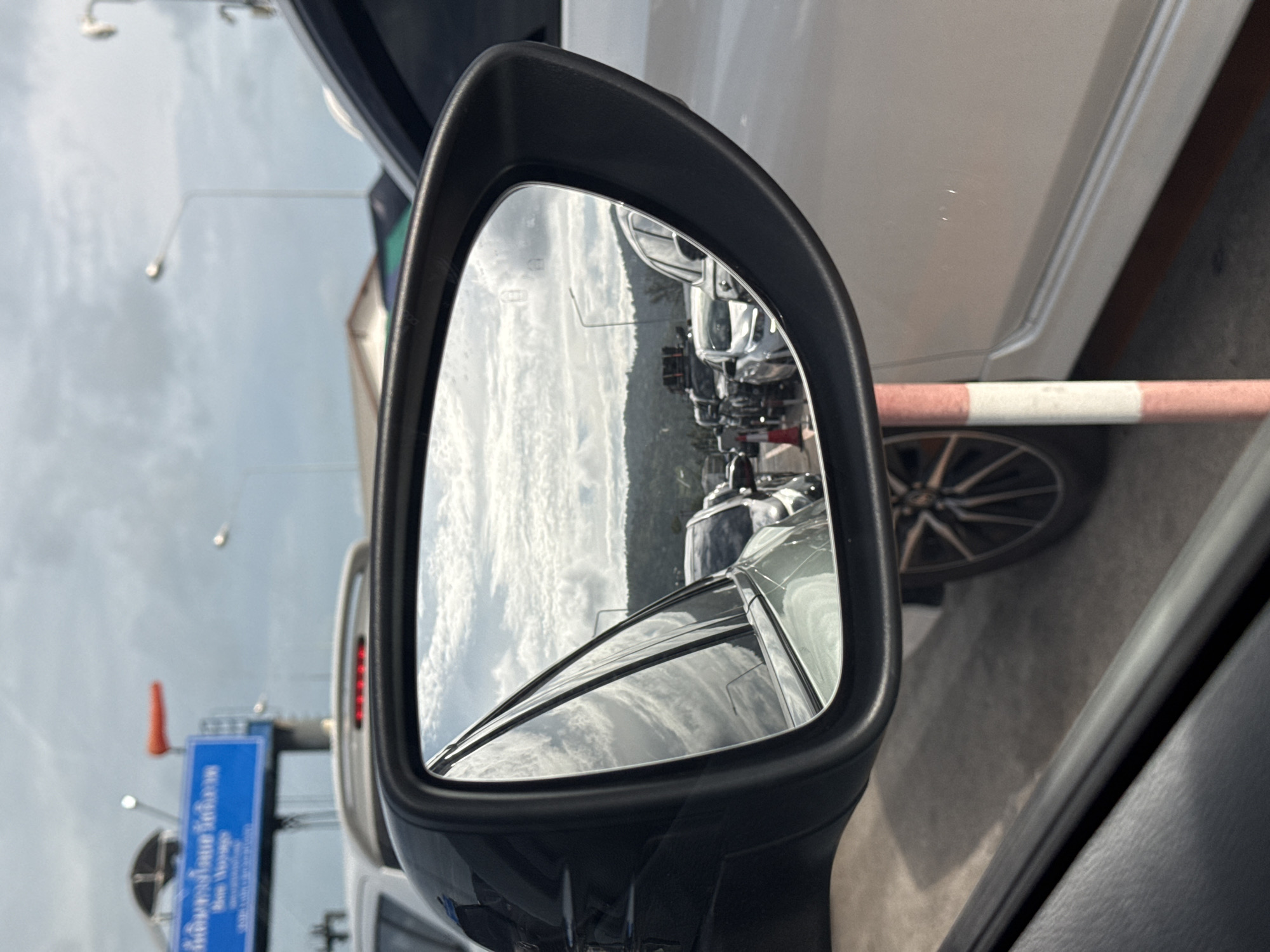 The image depicts a view from a vehicle in a traffic jam on the island of Koh Samui, Thailand, as seen through a car's side mirror. In the foreground, the black frame of the side mirror is visible, with the reflective surface showing a cloudy sky with scattered white clouds and distant green hills. A line of cars is also reflected in the mirror, with a silver car in the immediate reflection, followed by other vehicles. Outside the mirror, on the right side of the image, a portion of a white car is visible, along with a red and white striped traffic pole. In the background, above the cars, a blue sign with white lettering and an orange windsock is mounted on a pole. The sky is overcast with visible street lights. The scene suggests a typical day in a busy area on Koh Samui, with vehicular traffic and infrastructure elements common to the region.