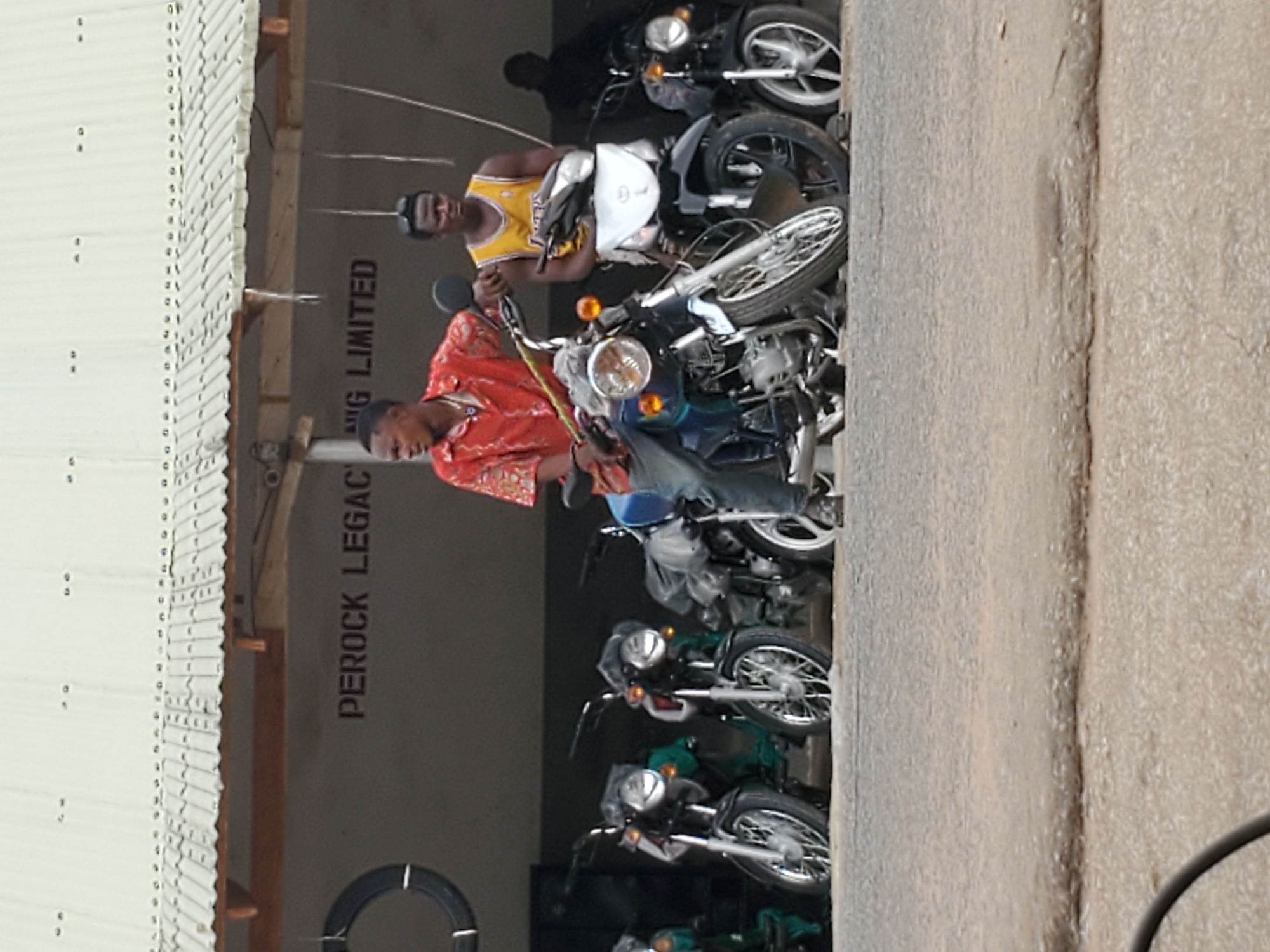 The scene depicts a commercial area in Mararaba, Nigeria, with "PEROCK LEGACY LIMITED" signage visible. Several motorcycles are parked in front of the building. Two men are seated on the motorcycles. One man wears a red patterned shirt, and the other wears a yellow and black Lakers jersey. Other motorcycles are parked nearby. The image is taken outdoors during daylight.