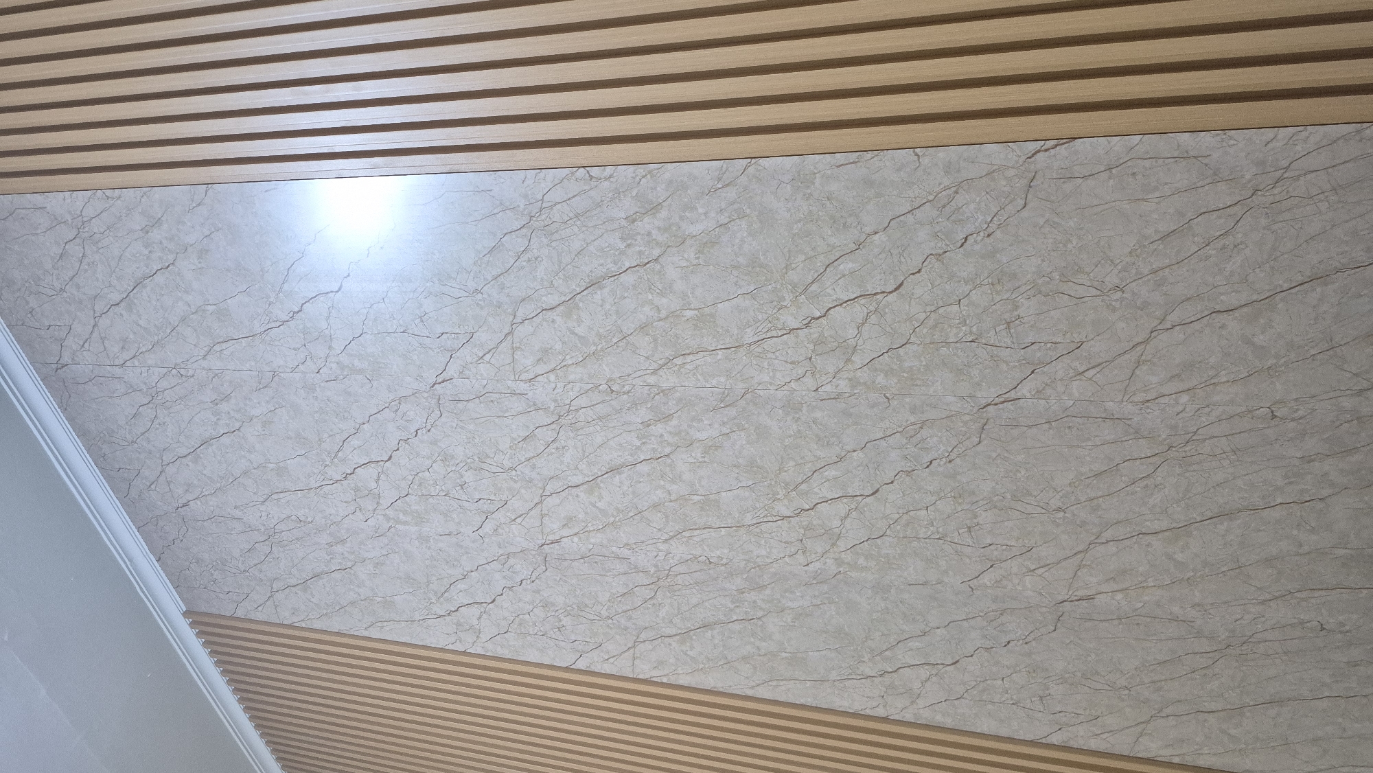 The image displays a wall with a decorative finish. The wall is divided into three sections. On the left and right, vertical wooden slats create a ribbed texture. The central section features a marble-patterned panel. The marble pattern is a light beige with darker brown veining, giving it a natural stone appearance. A bright light source reflects strongly off the central panel, suggesting a polished or glossy surface. The top edge of the wall meets a white ceiling, indicated by a white molding or cornice. The image is taken from a low angle, looking upwards towards the ceiling. No people, animals, or specific locations within South Tangerang, Indonesia are visible or indicated.
