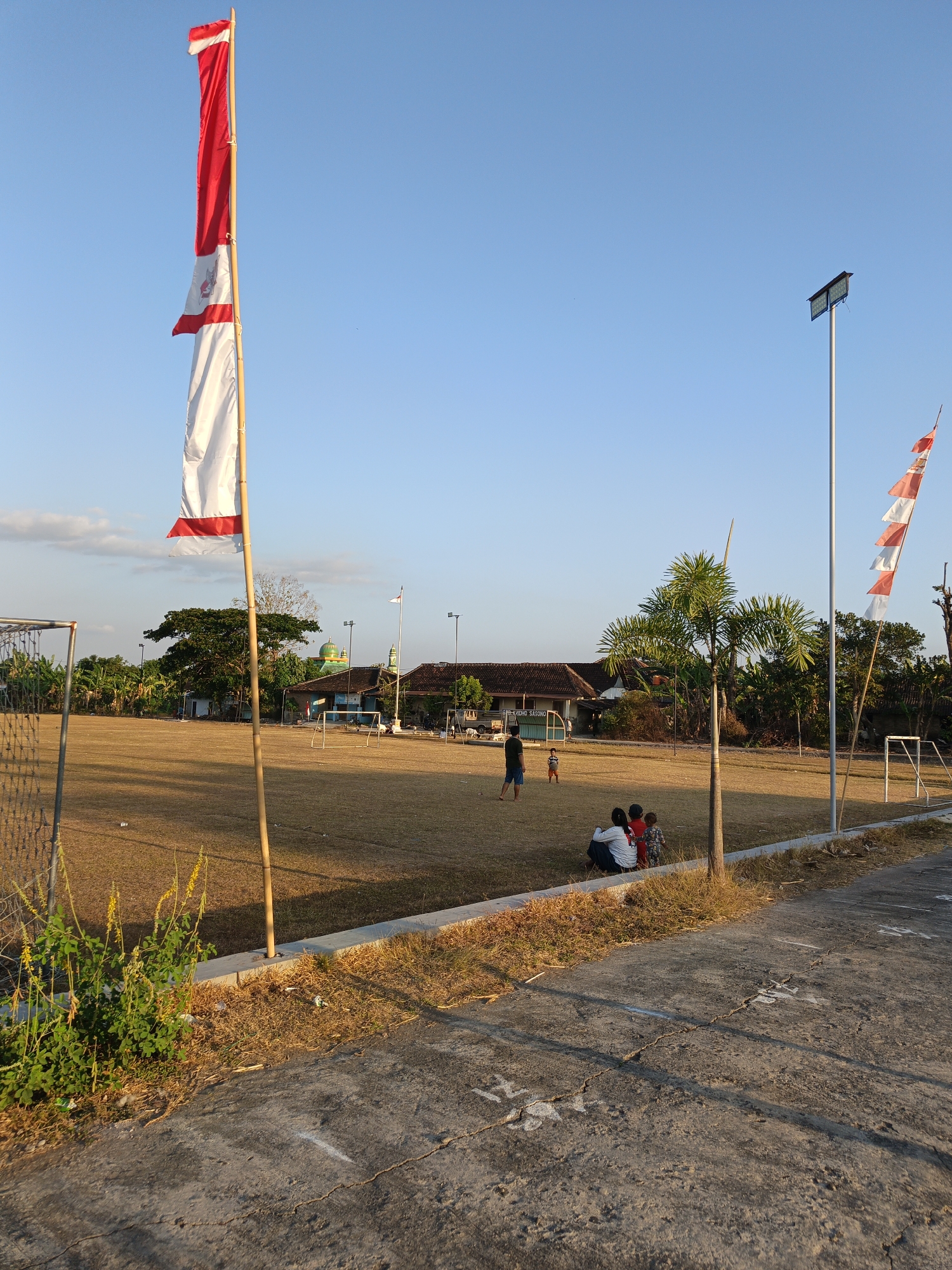 This image depicts an outdoor scene at what appears to be a community field or sports ground in Trucuk, Indonesia, with a clear blue sky. In the foreground, a concrete pathway runs diagonally across the lower portion of the frame. To the left, a patch of green vegetation with yellow flowers grows along a low concrete barrier that separates the pathway from the field. A chain-link fence is visible on the far left.
Dominating the left side of the image is a tall bamboo pole flying a red and white flag, which is partially unfurled and appears to be a national flag. Further into the field, several other flags are visible on poles, including a smaller red and white flag on the right.
The field itself is covered in dry, yellowish grass, indicating a sunny or dry season. There are several white markings on the field, suggesting it is used for sports like soccer, with goalposts visible in the background.
In the middle ground, a few people are present. On the far left, a man and a child are standing. Further to the right, a group of people, appearing to be a family with two children and an adult, are sitting on the grass, facing away from the camera. Behind them, a palm tree stands tall.
In the background, buildings with tiled roofs and a green dome, possibly a mosque, are visible, surrounded by trees. A tall lamppost with a solar panel is also present on the right side of the field. The overall atmosphere is one of a peaceful, community gathering in a rural or semi-urban setting.
