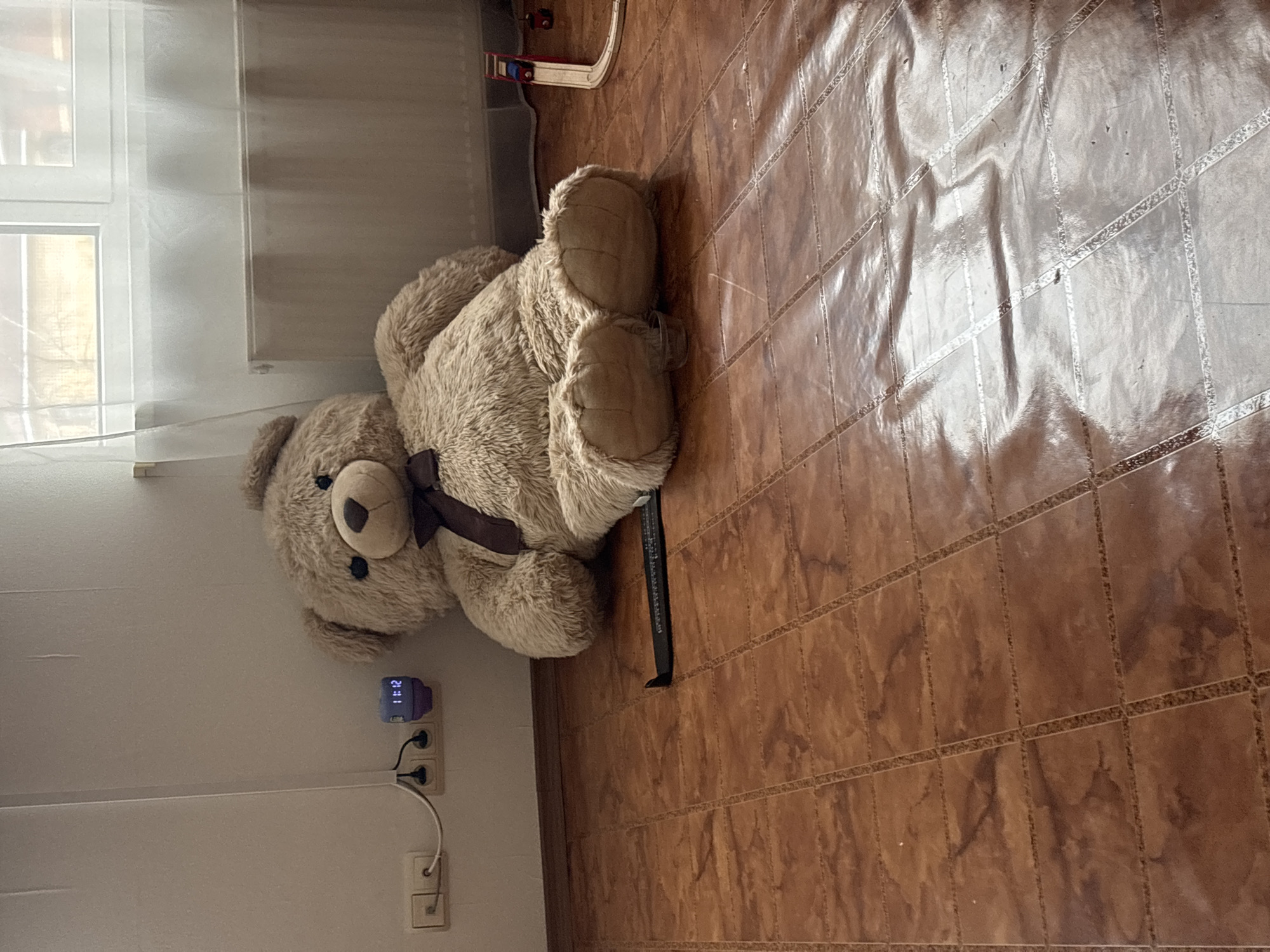 A tan teddy bear with a brown ribbon around its neck lies on its side on a tiled floor. A digital clock showing "21:12" is plugged into an electrical outlet on the wall next to the bear. The wall is white and has a window with white curtains to the left of the bear. In the background, there is a child's toy track. The location is Ферма, Russia.