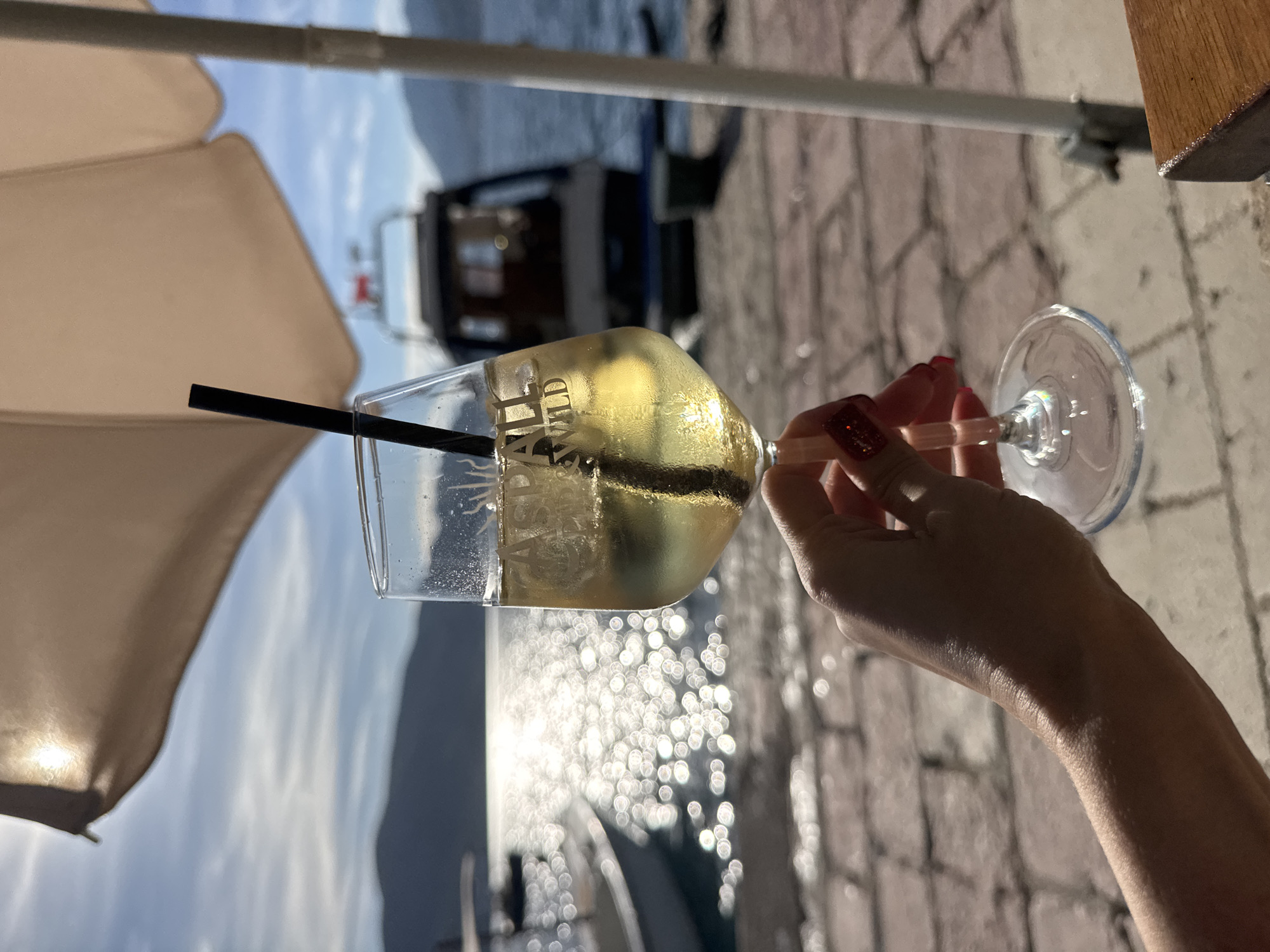 A close-up, eye-level shot captures a fair-skinned hand, featuring red glitter nail polish, holding a stemmed wine glass. The glass contains a pale yellow liquid with ice cubes and a black straw, and displays a partially visible white logo that reads "ASPA WILD." The hand and glass are positioned in the shade provided by a large, light-colored umbrella, a portion of which is visible in the upper frame and strongly illuminated by sunlight on its left side. A white vertical pole, likely part of the umbrella stand, is visible on the right.
The setting is outdoors, on a paved surface composed of light-colored interlocking bricks or stones. Beyond the paved area, a body of water shimmers with intense sunlight reflections, creating a bokeh effect. A white boat is partially visible on the left side of the water, and a larger, dark-hulled boat with a cabin is docked further out on the right. Distant mountains or hills line the horizon under a clear blue sky with scattered white clouds. A wooden surface, possibly a table edge, is visible at the bottom right of the frame. The overall impression is a sunny, waterfront location during daytime.